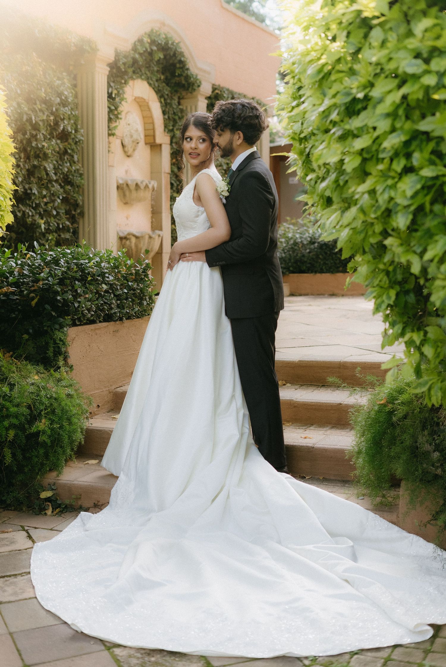 A wedding couple poses on stone steps against a Mediterranean-style wall with trailing vines.