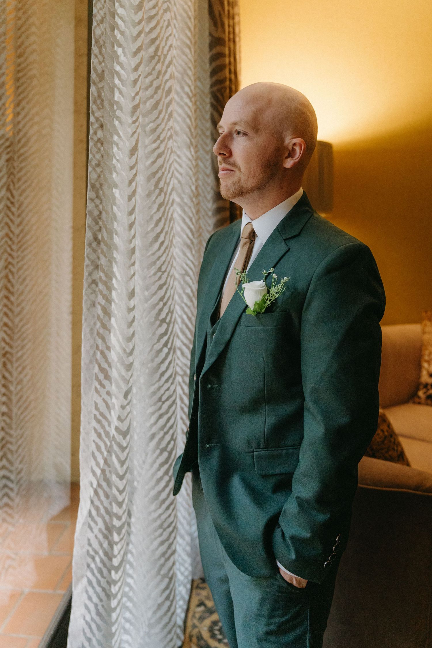 A groom in an elegant emerald green suit and boutonniere stands thoughtfully by a window before a wedding ceremony.