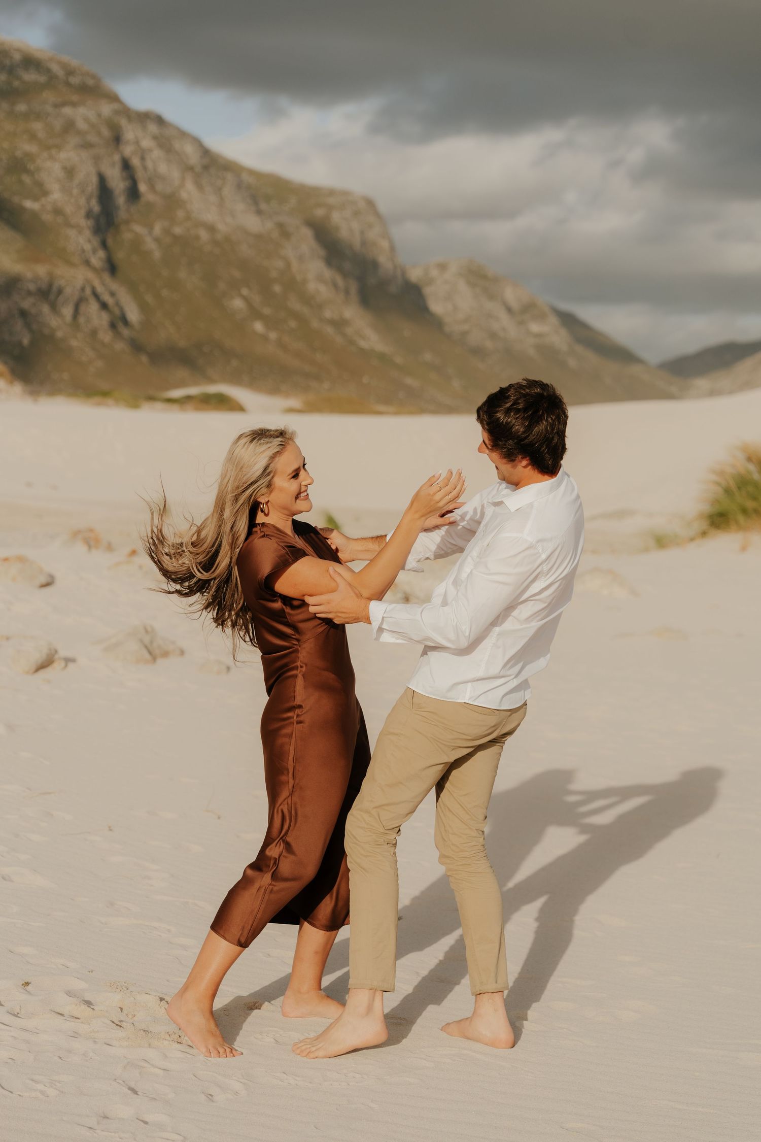 Couple dancing barefoot on white sand dunes with mountains in background, woman in brown satin dress, man in white shirt.