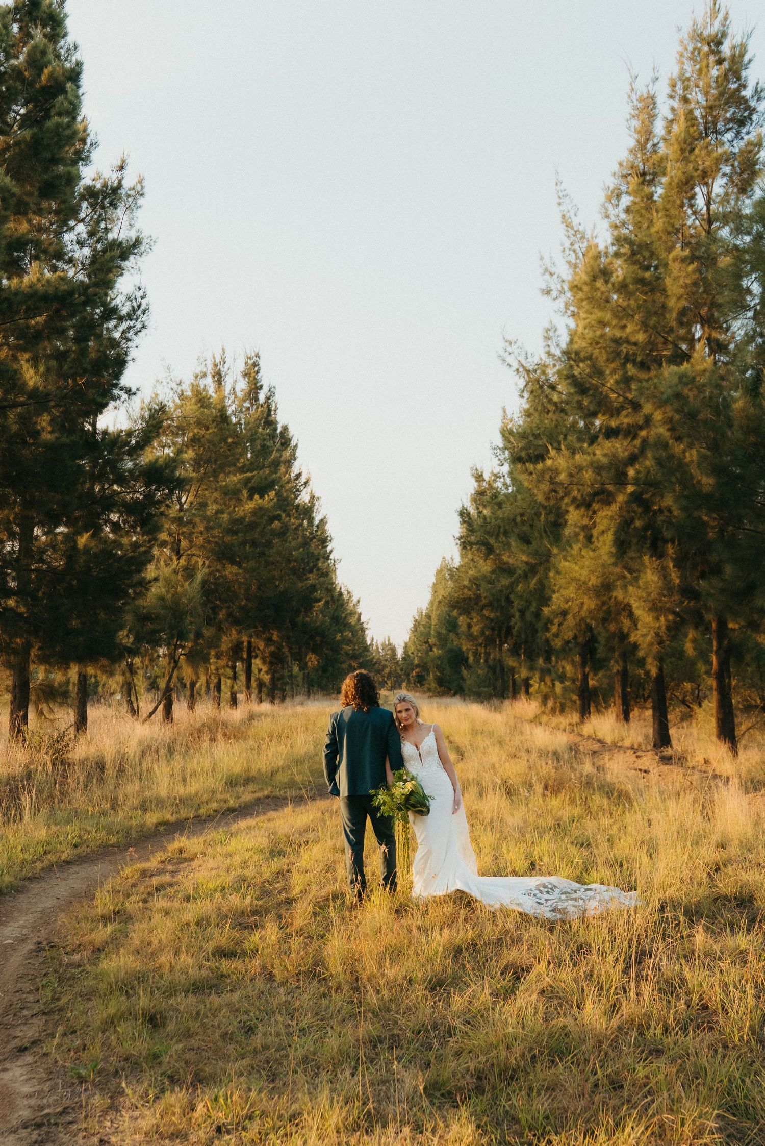 Bride and groom walk hand-in-hand down a tree-lined path at Rosemary Hill Farm in Pretoria in golden hour light.