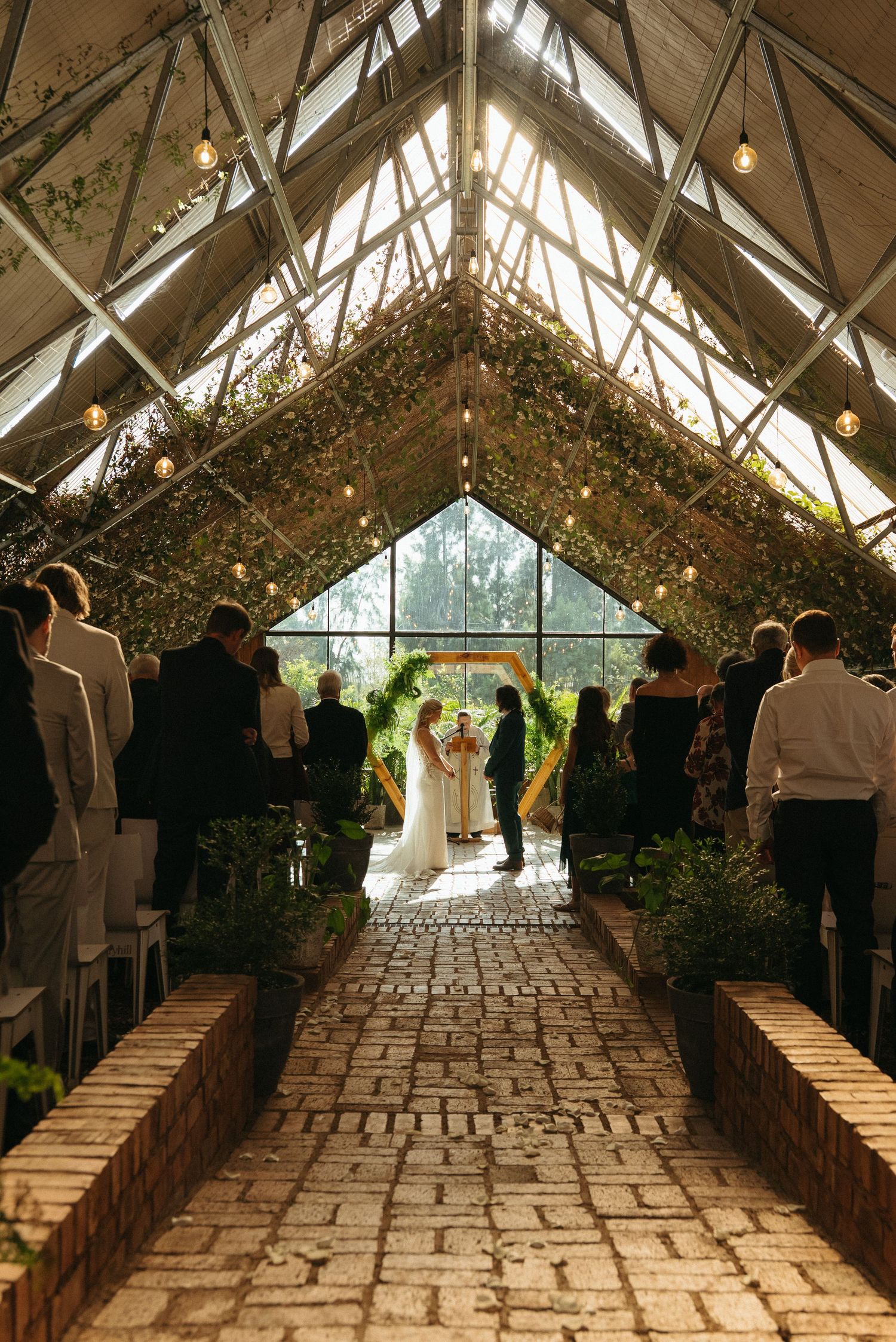 Wedding ceremony in a sunlit greenhouse with brick aisle, lush greenery, string lights, and guests seated on both sides.