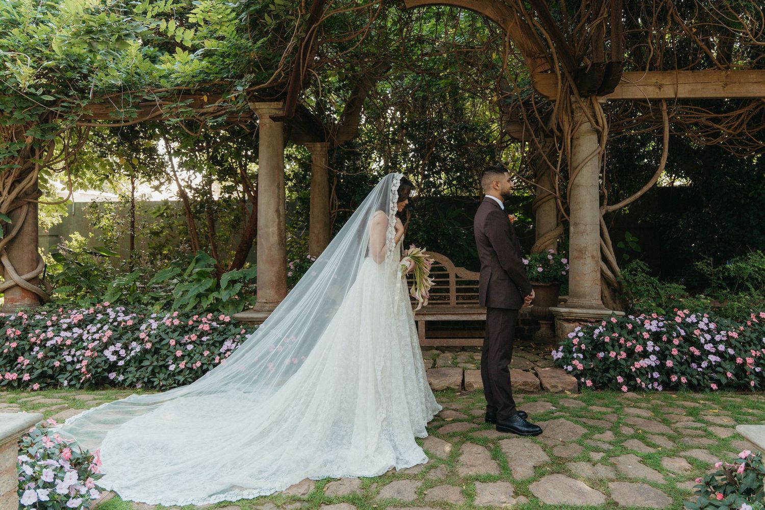 Bride in flowing white gown and cathedral veil stands with groom under floral garden arbor during outdoor wedding ceremony.