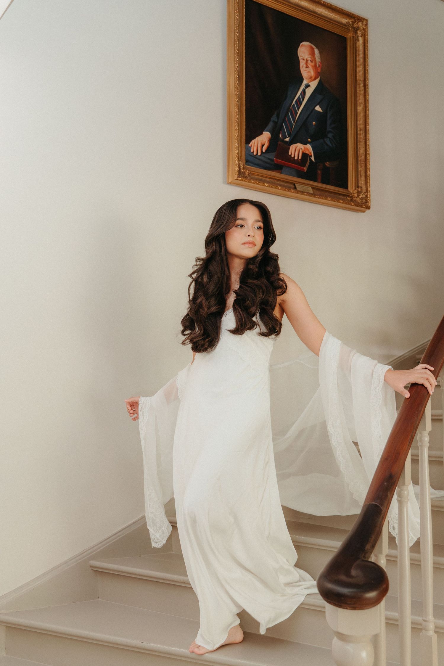 Young woman in flowing white gown descends elegant staircase beneath a formal portrait painting.