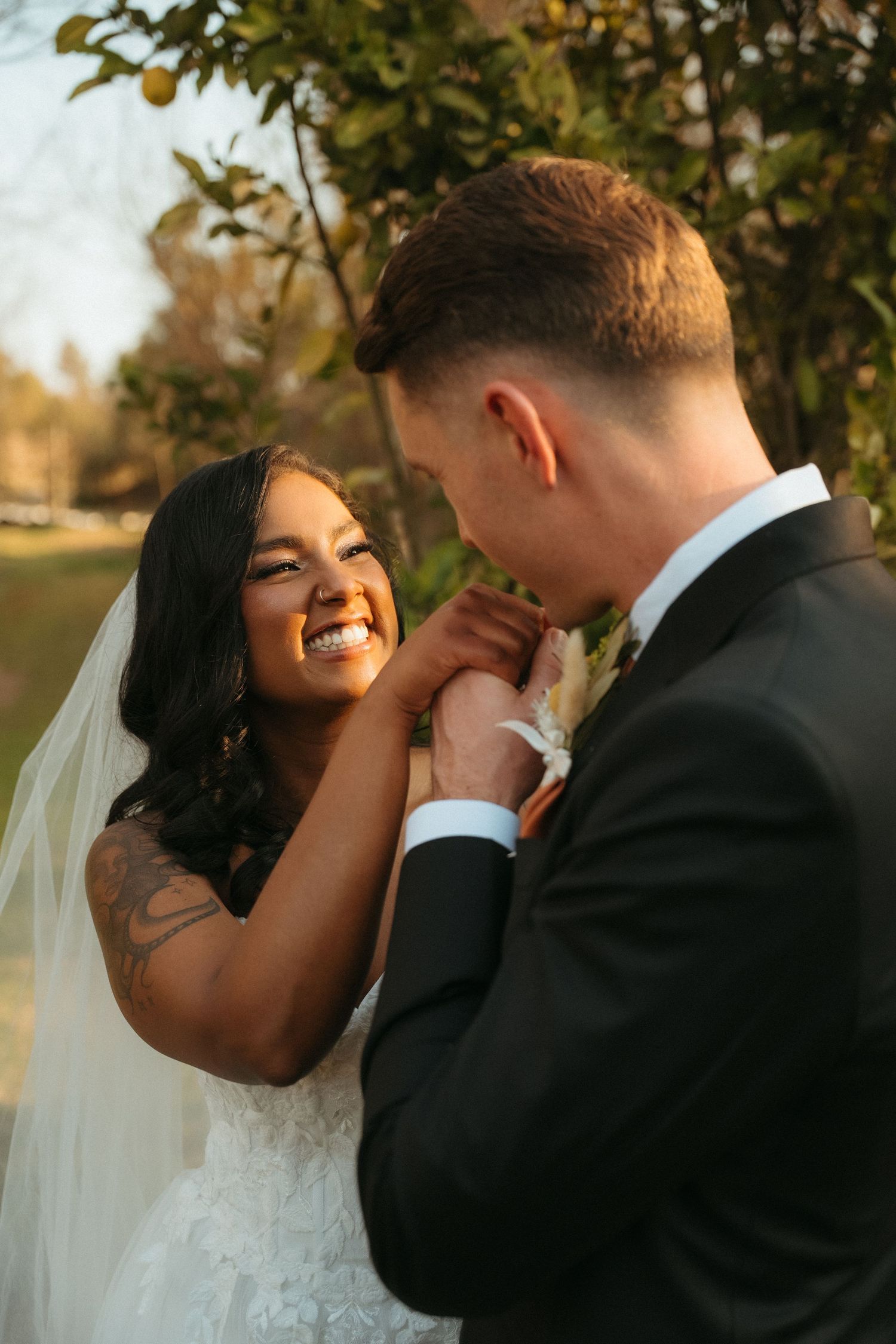 Smiling bride in a lace wedding gown and veil holds her groom's hand as he leans in, in golden outdoor light.