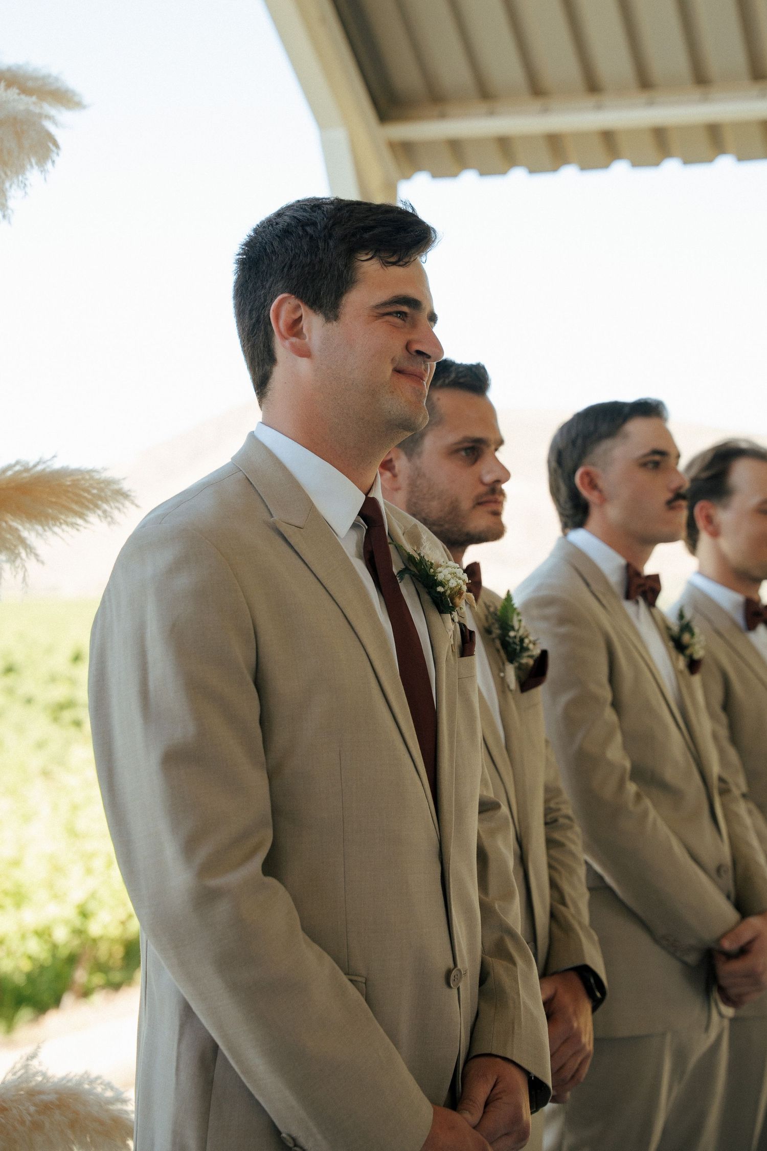 Groom in a tan suit and burgundy tie smiles at the altar beside groomsmen in matching suits with floral boutonnieres at an outdoor wedding.