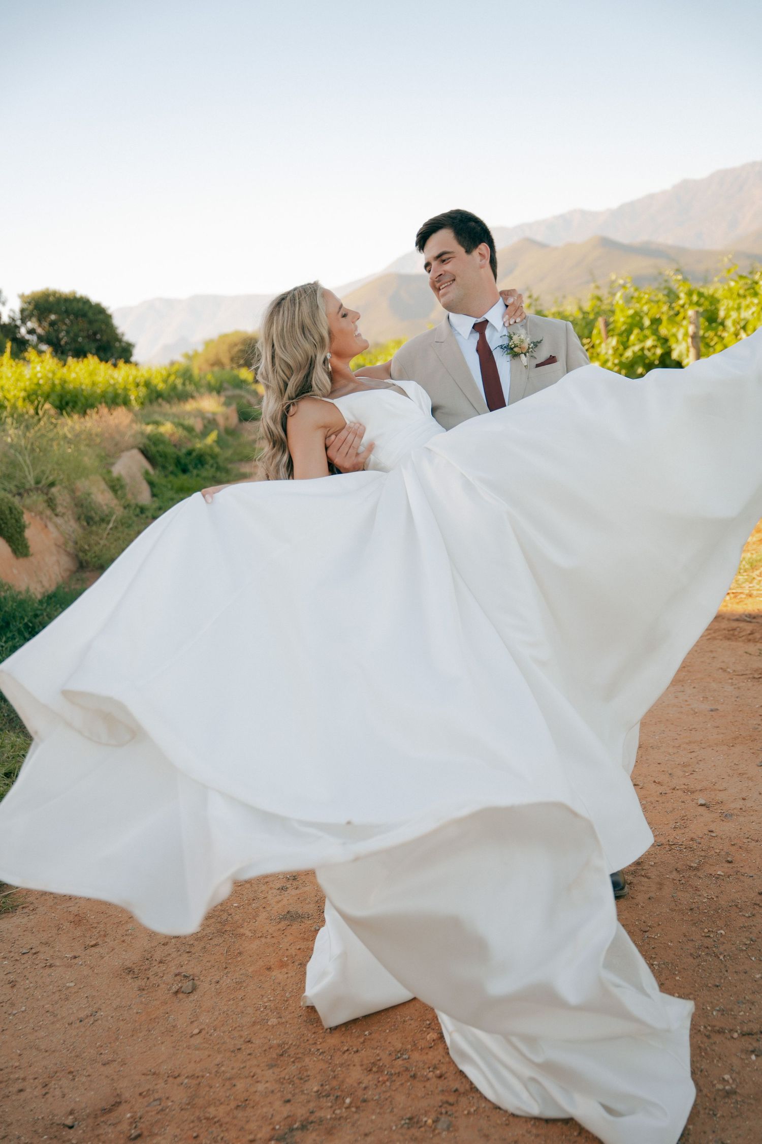 Groom in a tan suit lifts and twirls his smiling bride in a flowing white gown on a dirt path by vineyards with mountains behind.