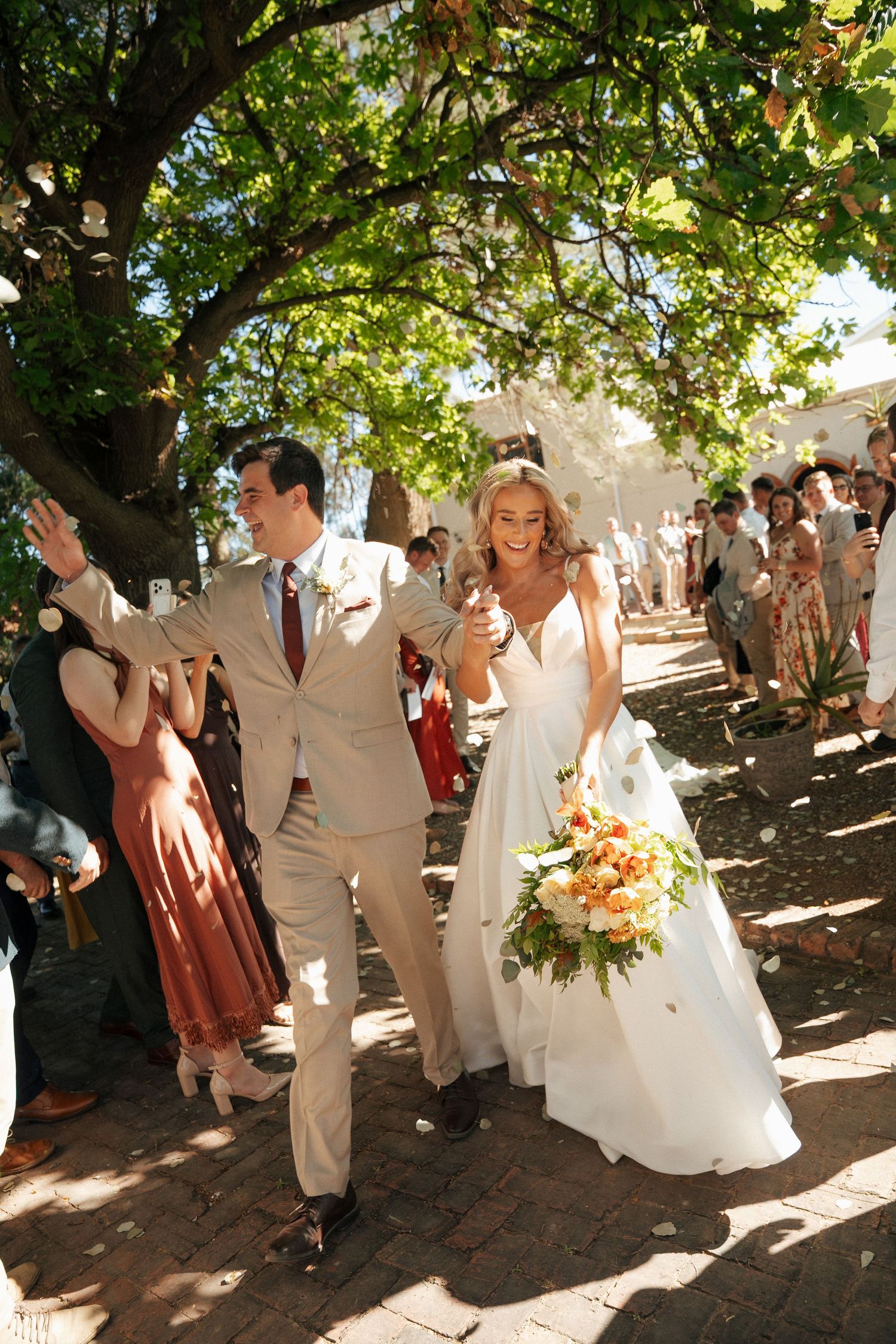 Smiling newlyweds walk under sunlit trees as wedding guests cheer, the bride holding a peach and white bouquet at Duvon Wine Estate.