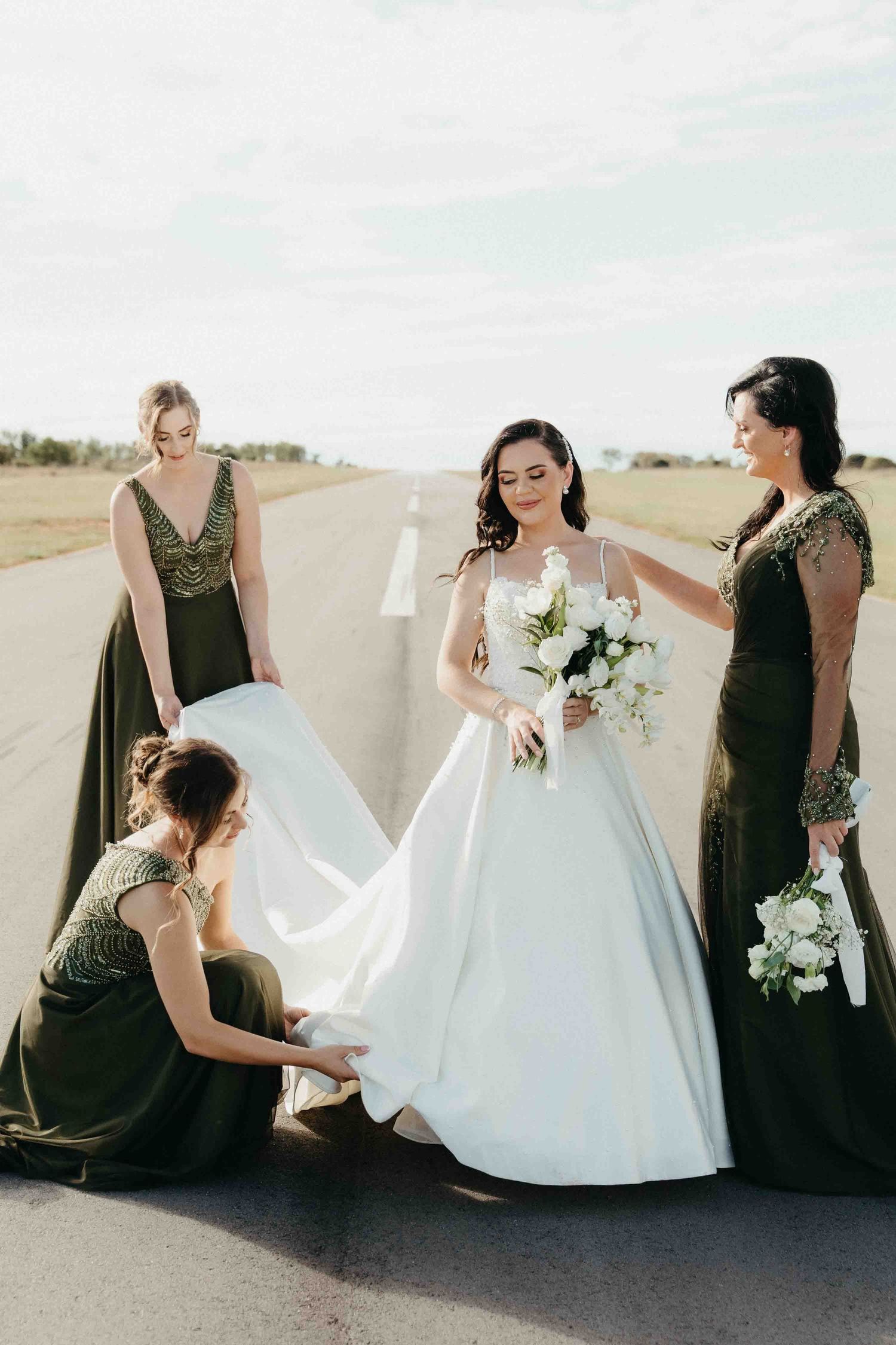Bride in white gown holds bouquet while three bridesmaids in dark green dresses adjust her train on an outdoor runway.