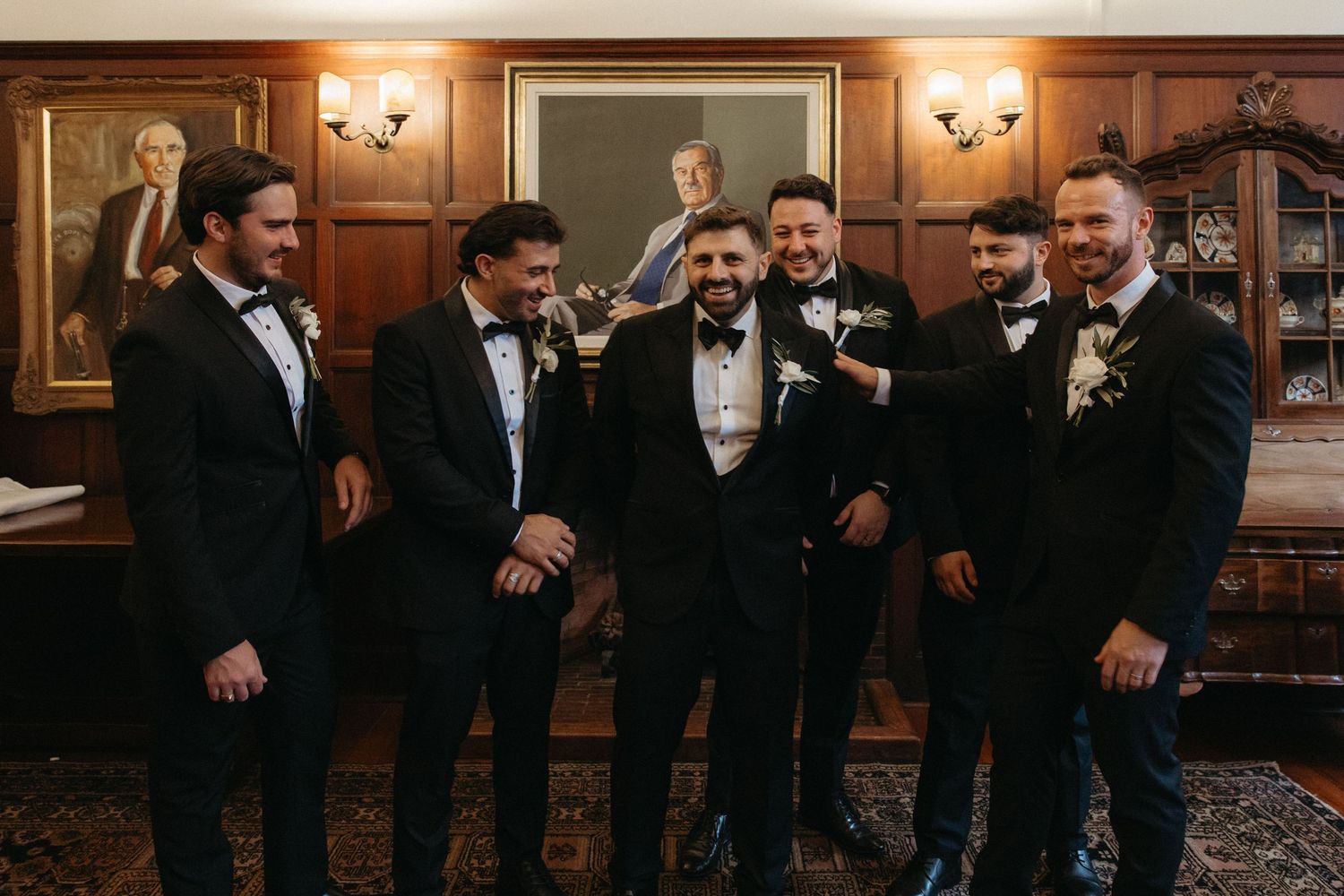 Six groomsmen in black tuxedos with bow ties and boutonnieres laughing together in an elegant wood-paneled room at Glenshiel