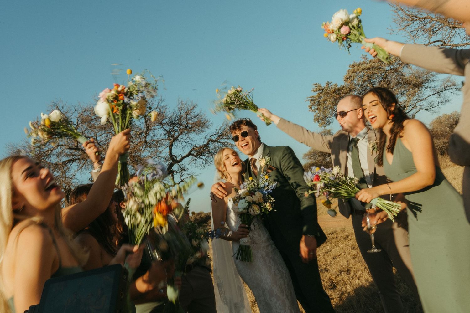Wedding party celebrating outdoors, bridesmaids in sage green dresses raising bouquets with joyful couple under clear blue bushveld sky.