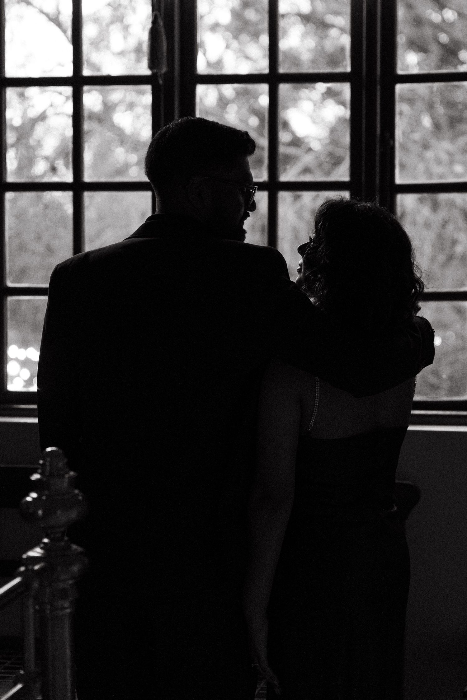 Black and white silhouette of a romantic couple embracing near a large window with trees visible outside.