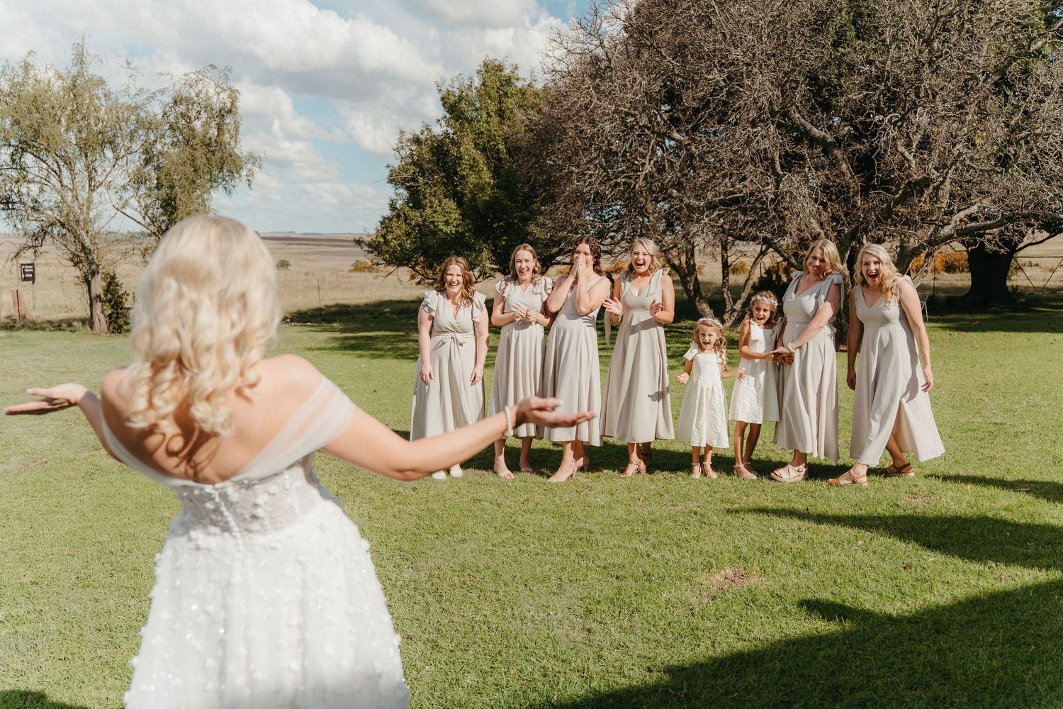 Bride in white gown faces her bridesmaids in silver dresses on a green lawn, as they react with joy and excitement.