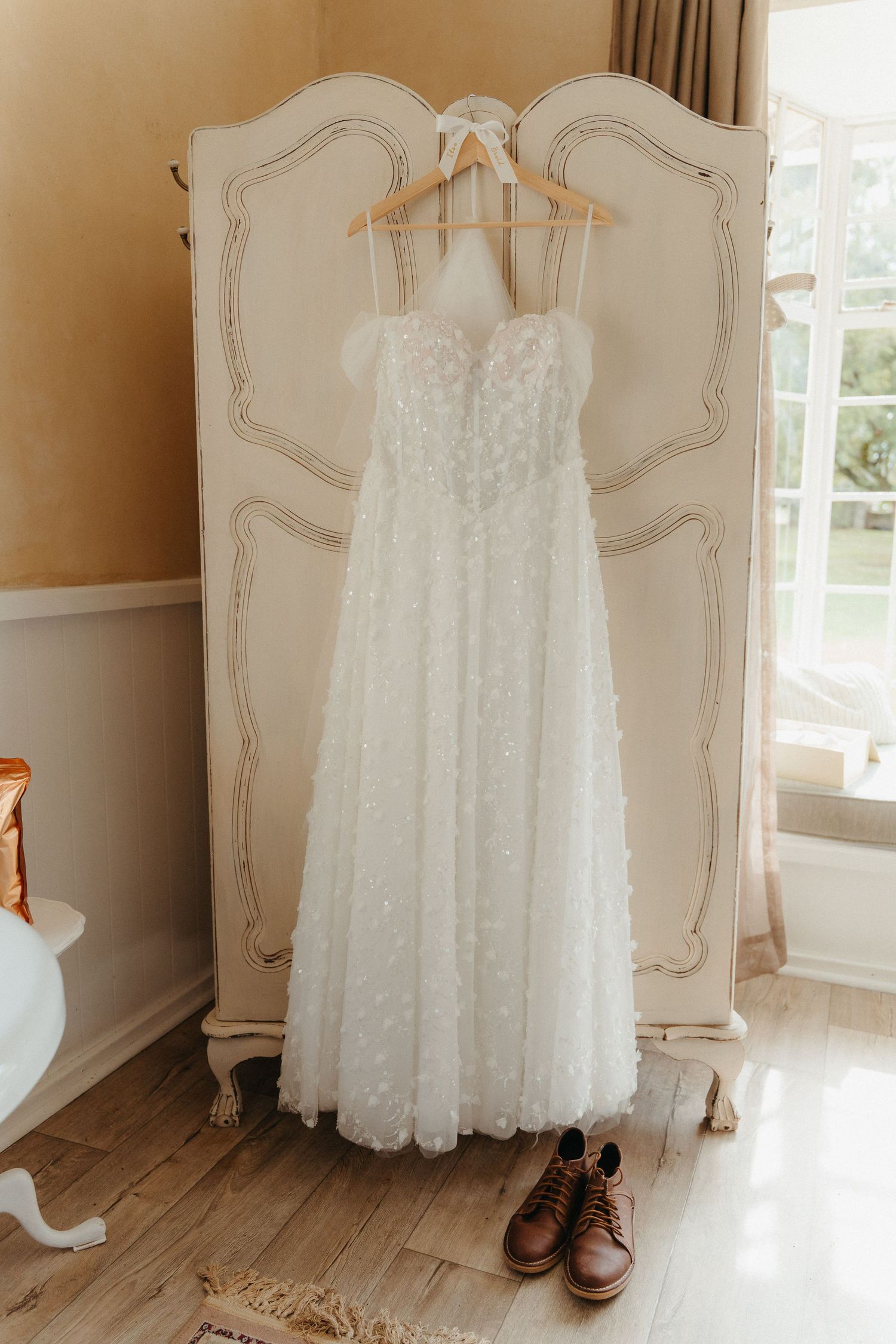 White floral lace wedding dress hanging on vintage cream armoire with brown leather boots below near bright window.