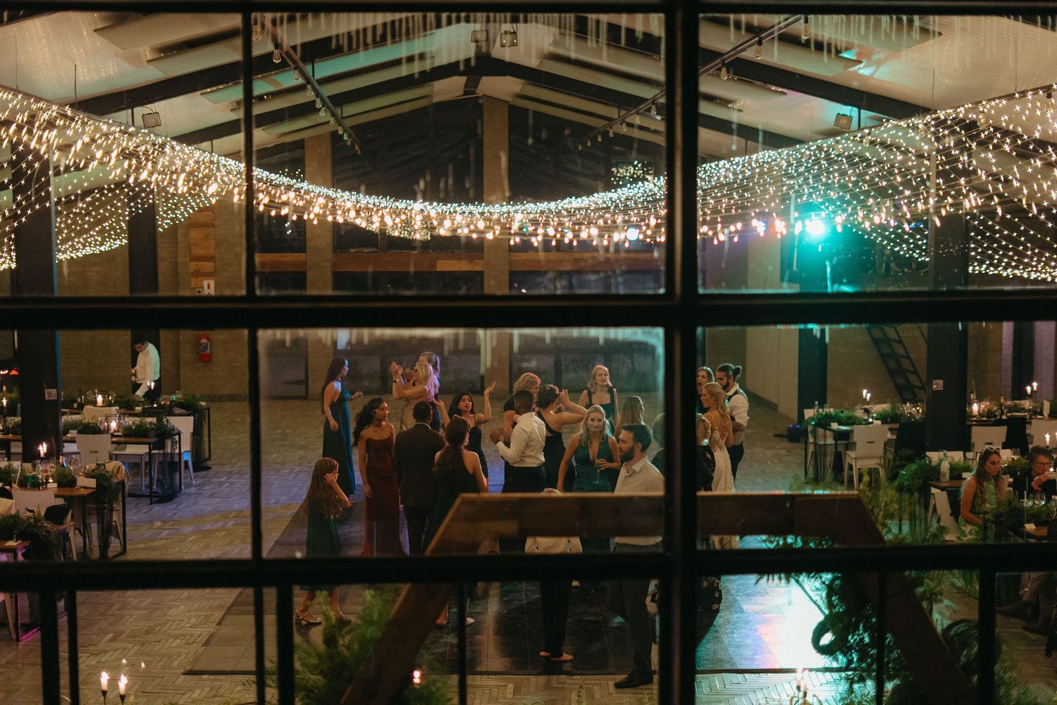 Elegant indoor wedding reception with string lights and guests dancing, viewed through large windows at night.