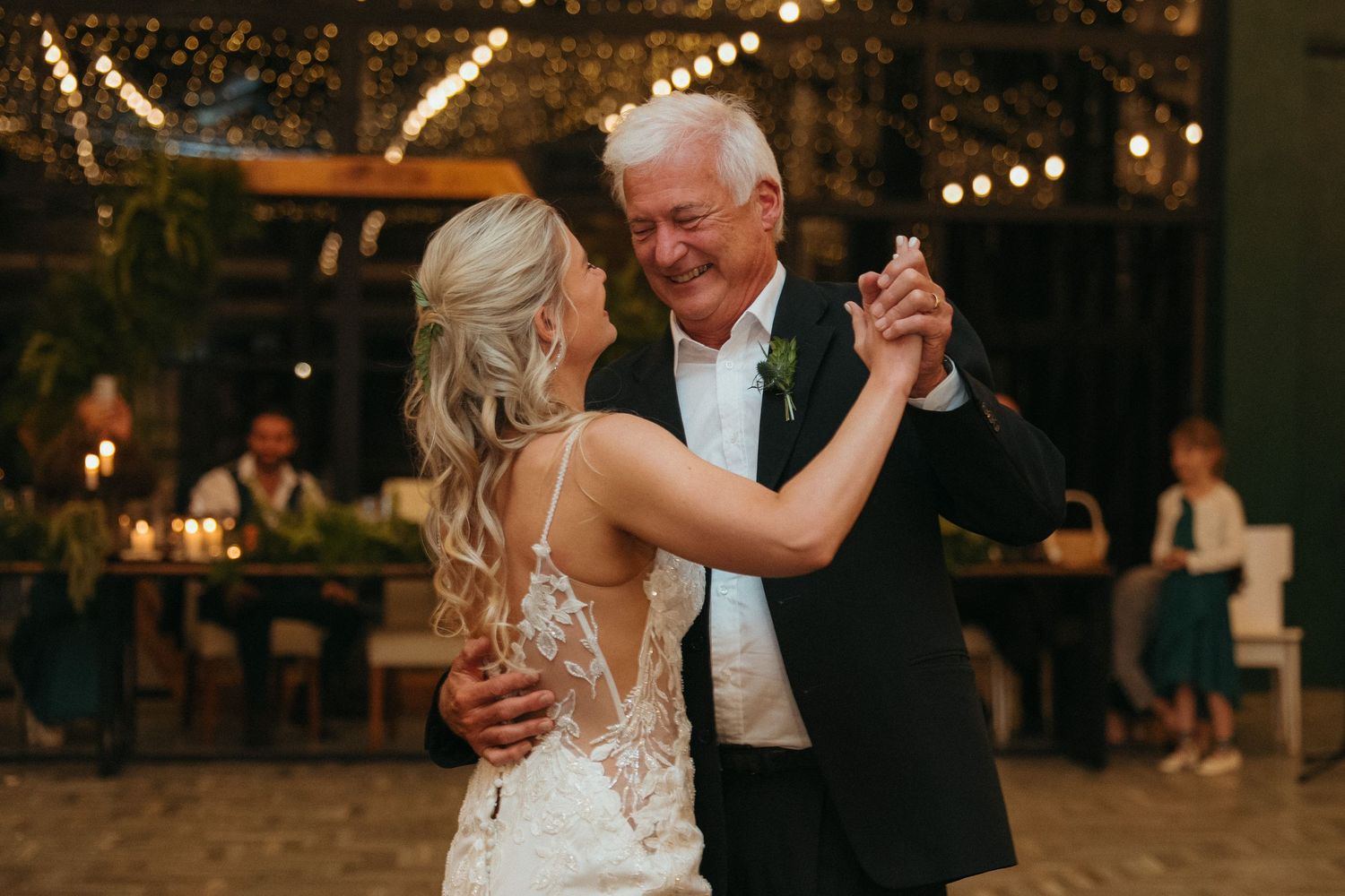 Bride and her father share an emotional first dance at a candlelit wedding reception with string lights overhead.