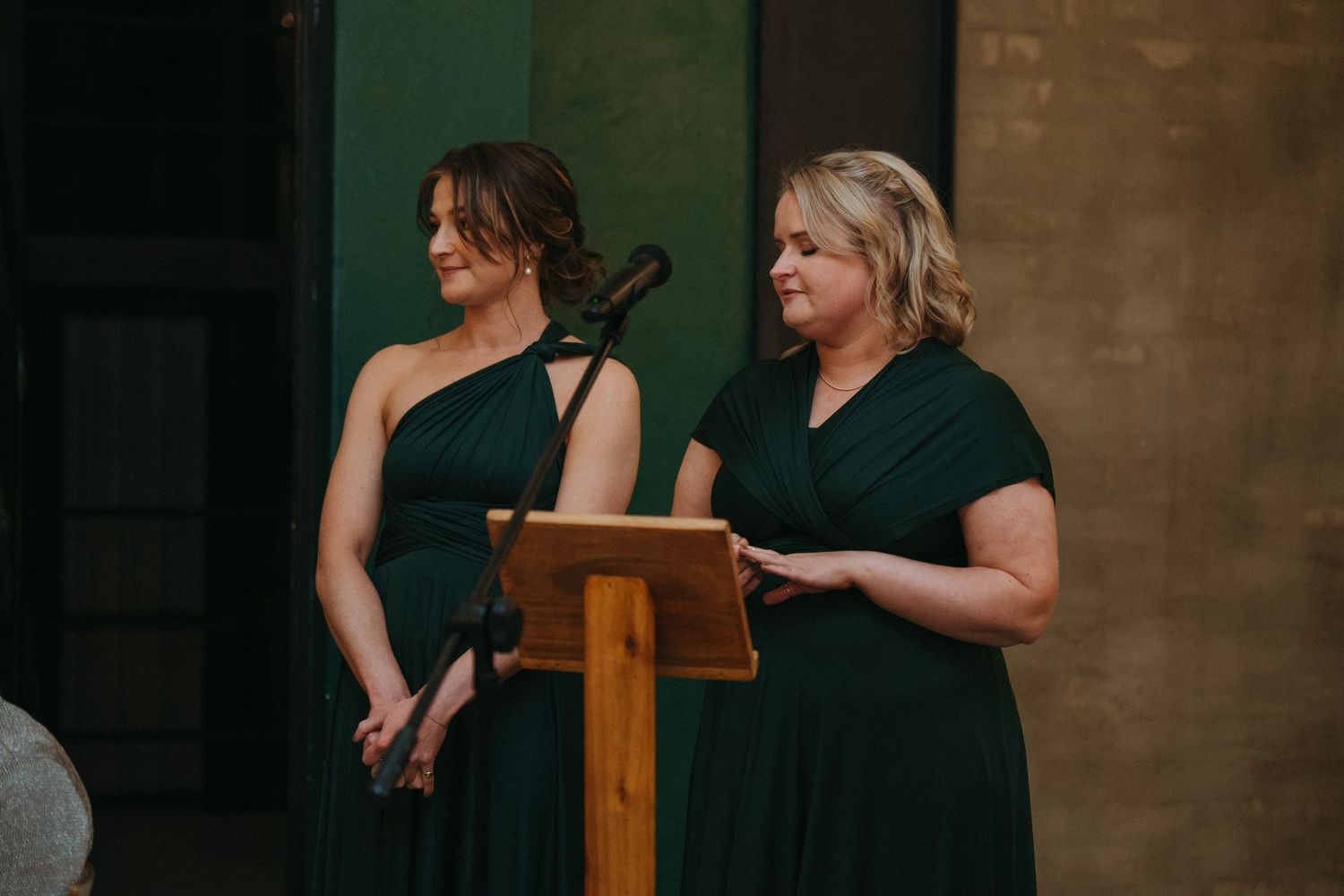 Two bridesmaids in elegant green one-shoulder dresses standing at a wooden podium during a wedding ceremony.