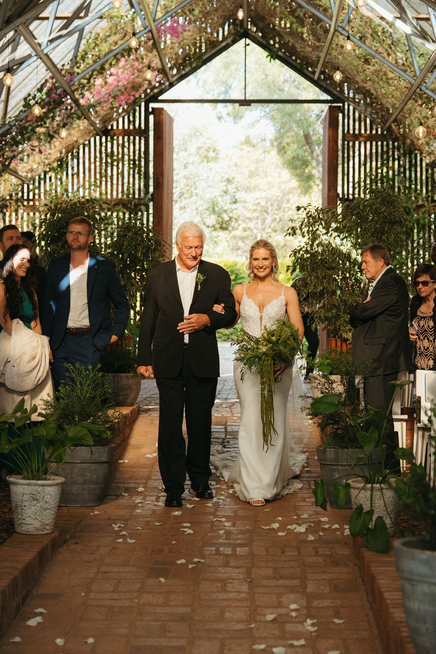 Bride walks down greenhouse aisle with older gentleman, holding greenery bouquet, rose petals on brick path.