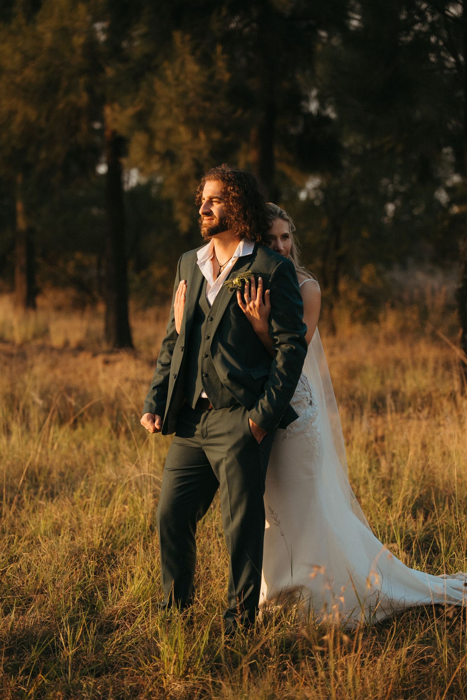 Bride in white gown embraces groom in dark suit standing in golden field at sunset with trees behind.
