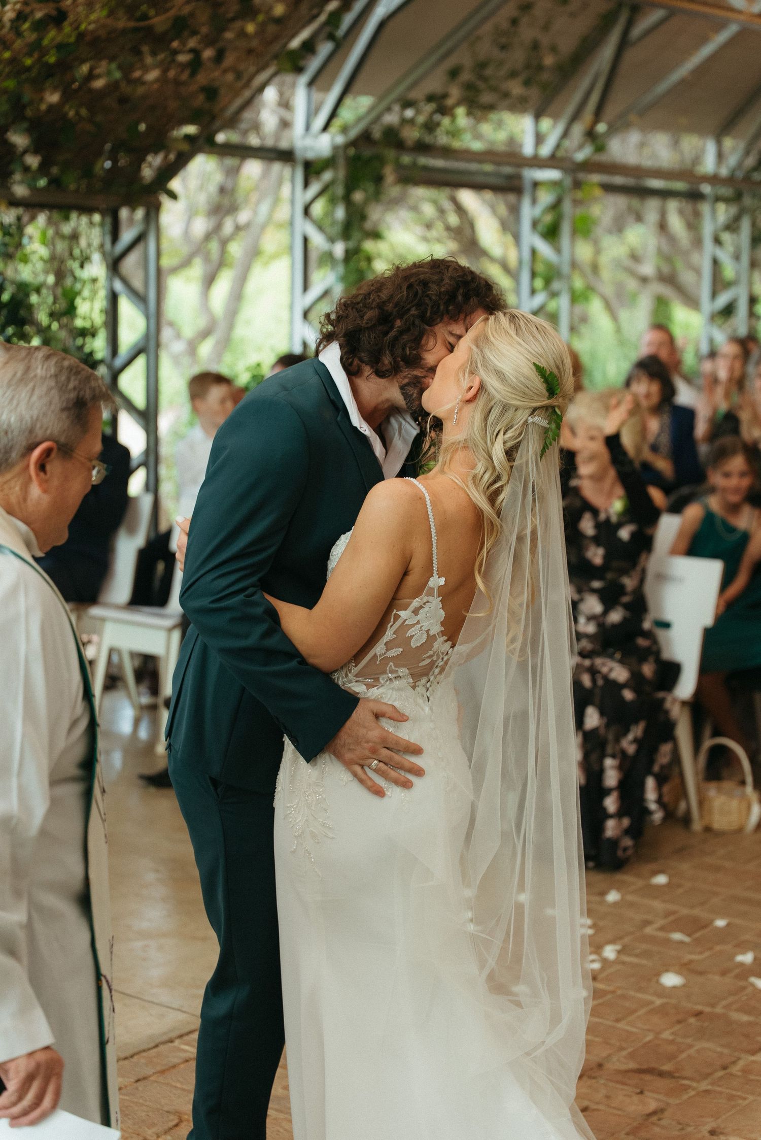 Bride and groom share their first kiss at an outdoor wedding ceremony, guests seated in background.