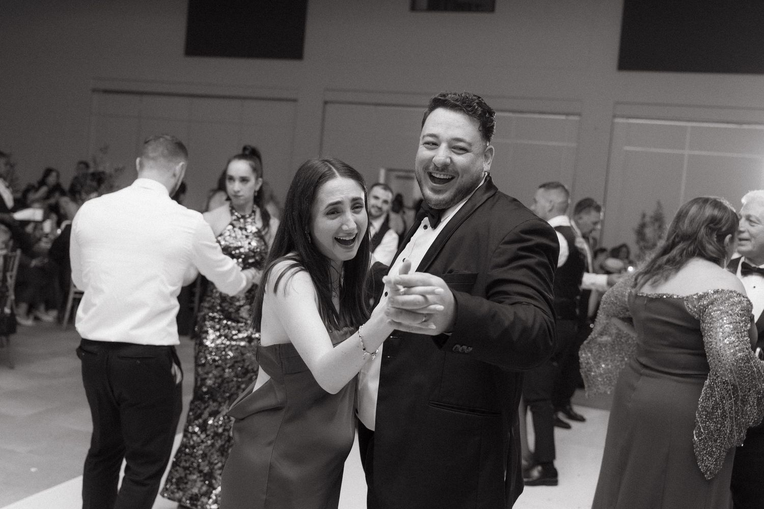 Black and white photo of a smiling couple dancing at a formal event surrounded by elegantly dressed guests.