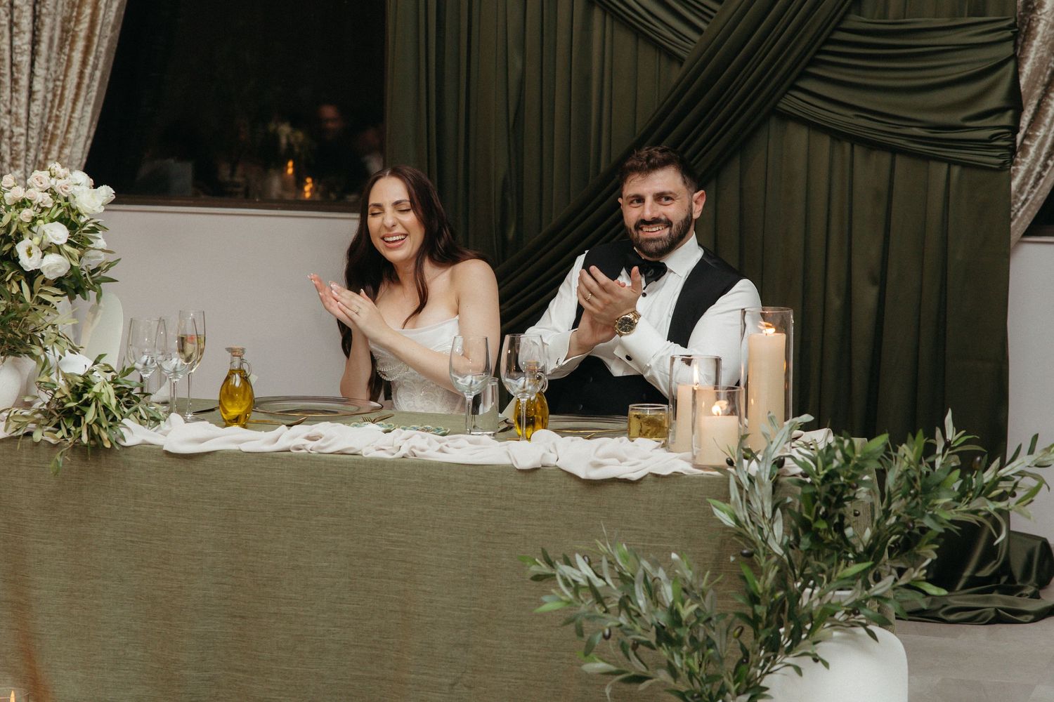 Bride and groom clapping at wedding reception table with candles, flowers, and green drapes.