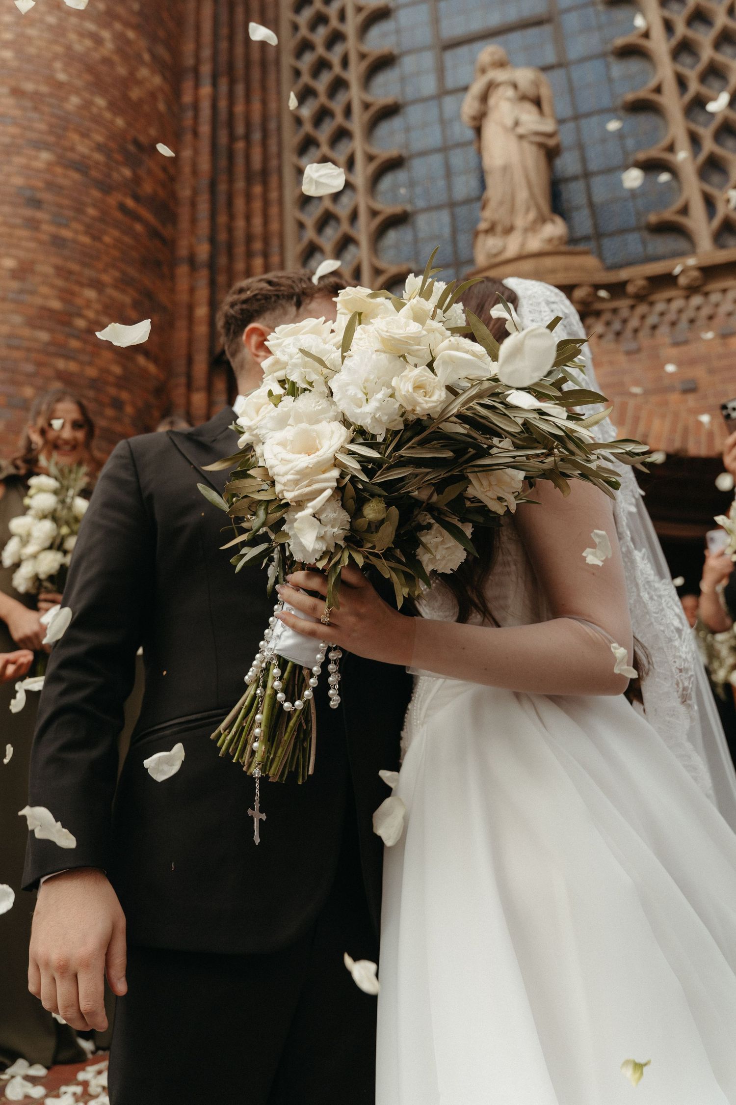 Bride and groom kissing behind white rose bouquet outside Rosebank Catholic Church with flower petals falling.