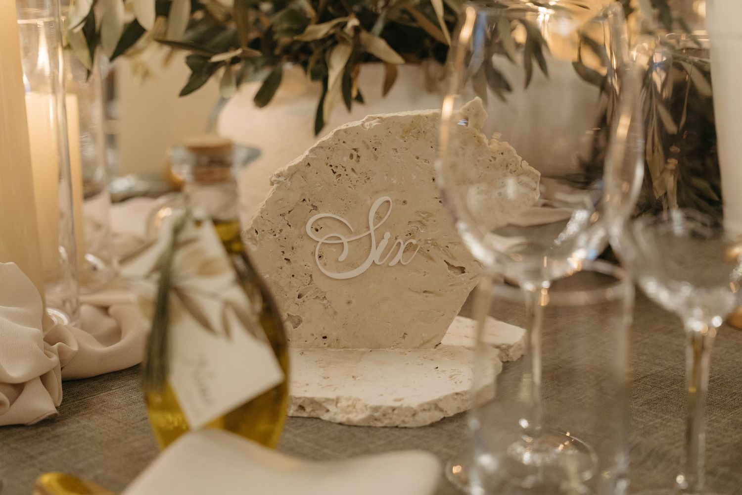 Elegant wedding table setting with engraved stone place card, olive oil bottle, wine glasses, and candles.