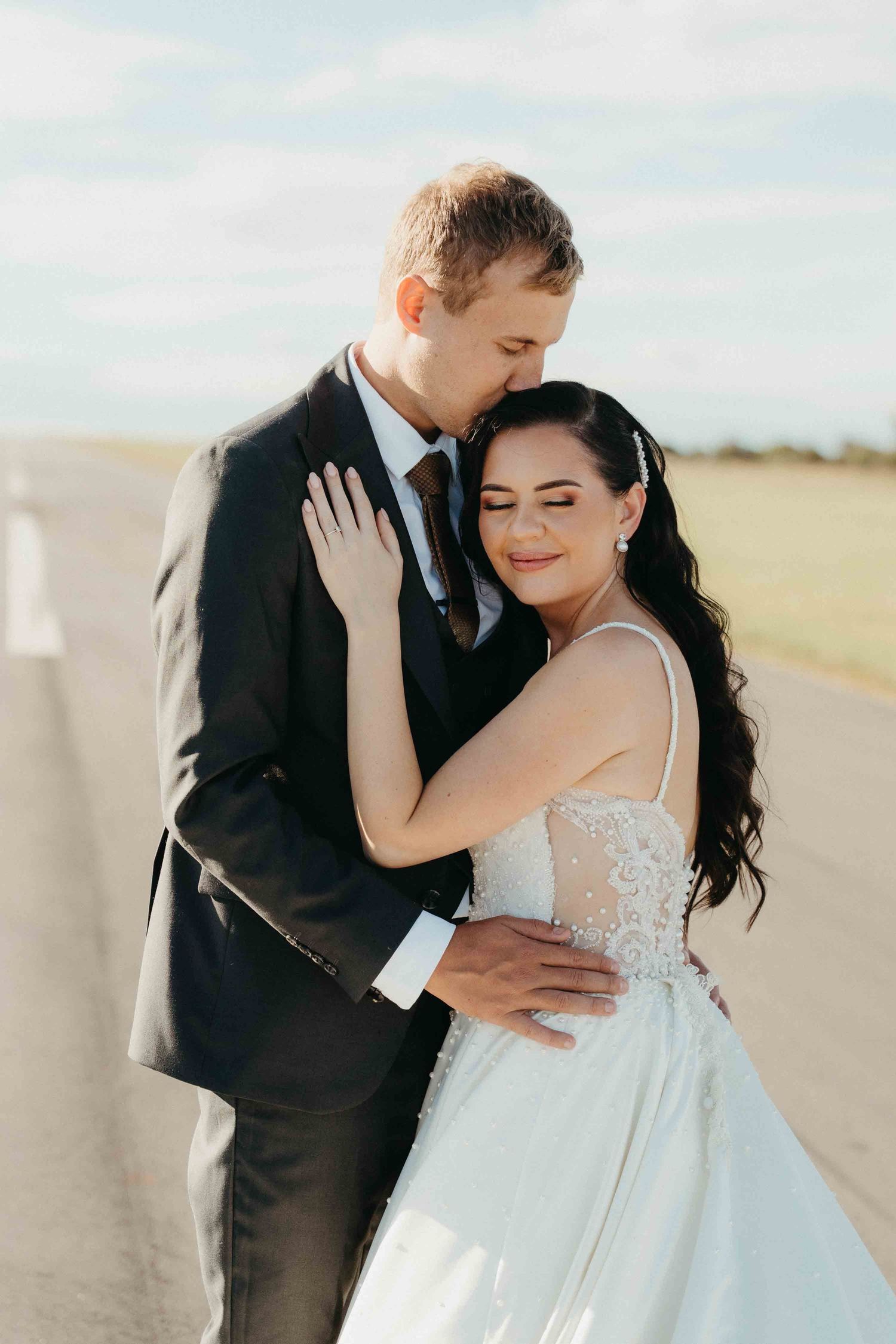 A bride and groom share a romantic embrace on an open road, dressed in a white gown and dark suit on their wedding day.