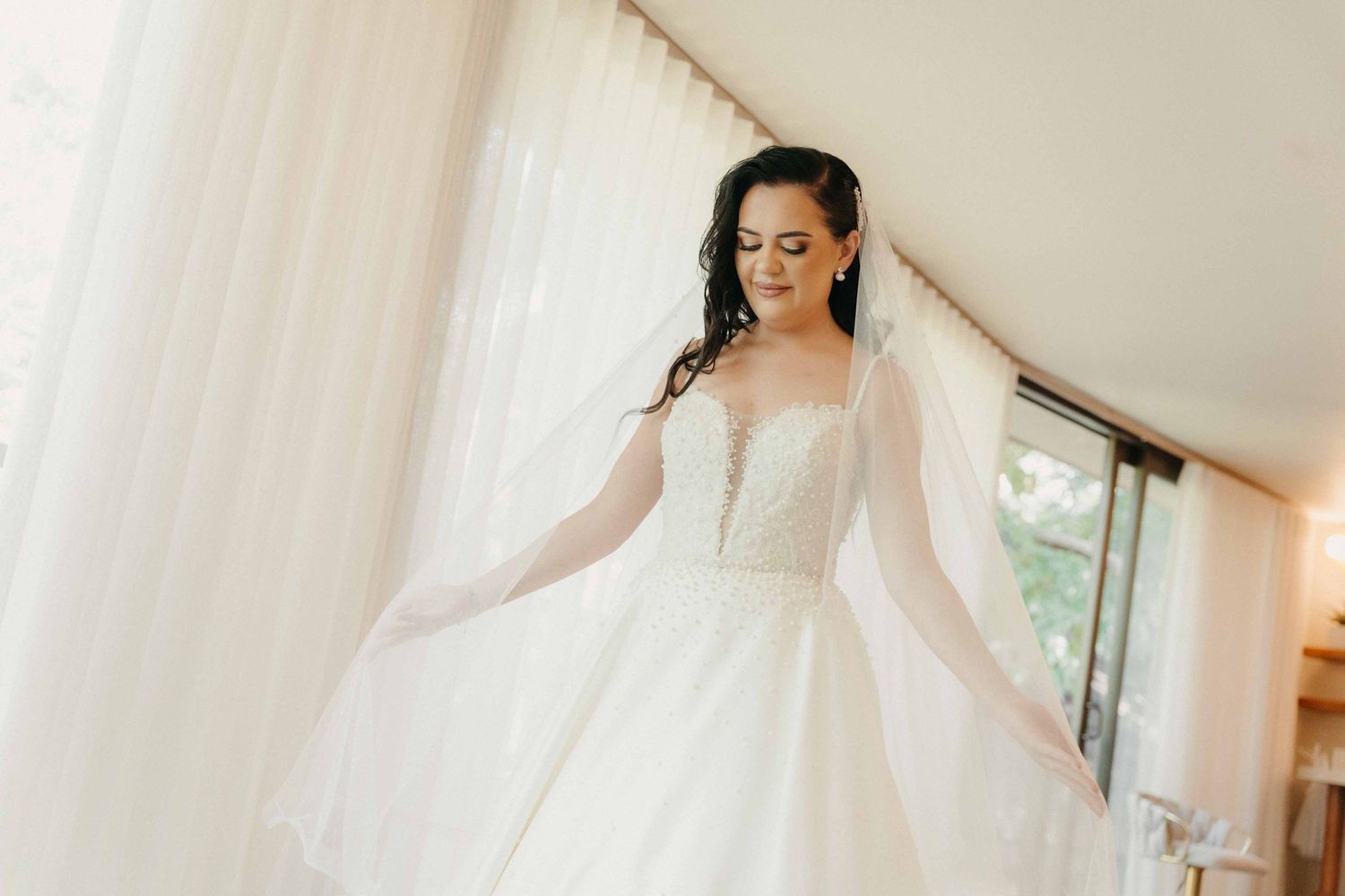 Bride in elegant white ball gown with sheer sleeves holding flowing veil near curtained window on wedding day.