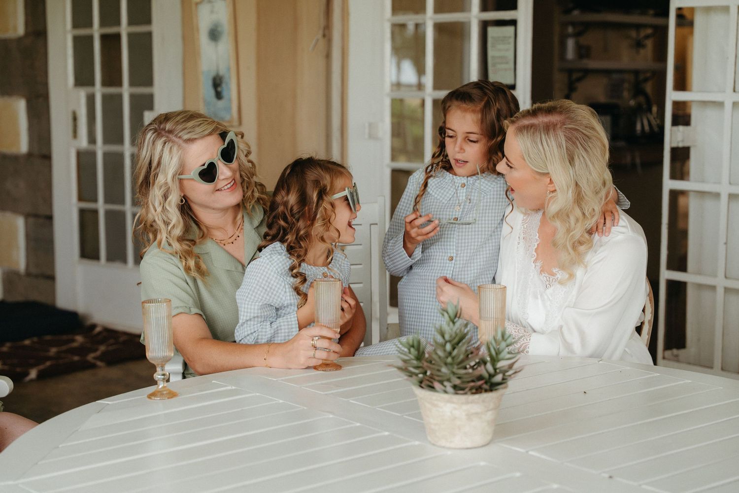 Two women and two young girls sit together at a white outdoor table, sharing a warm family moment on a porch.