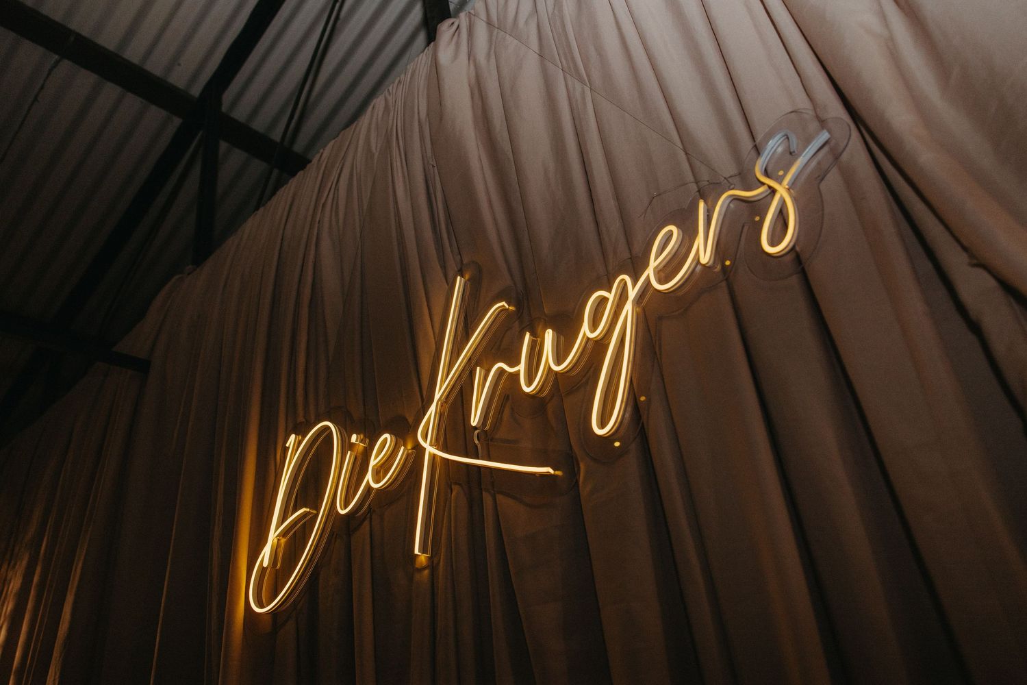 Golden neon sign reading Die Krugers in cursive script, mounted against draped beige fabric backdrop.
