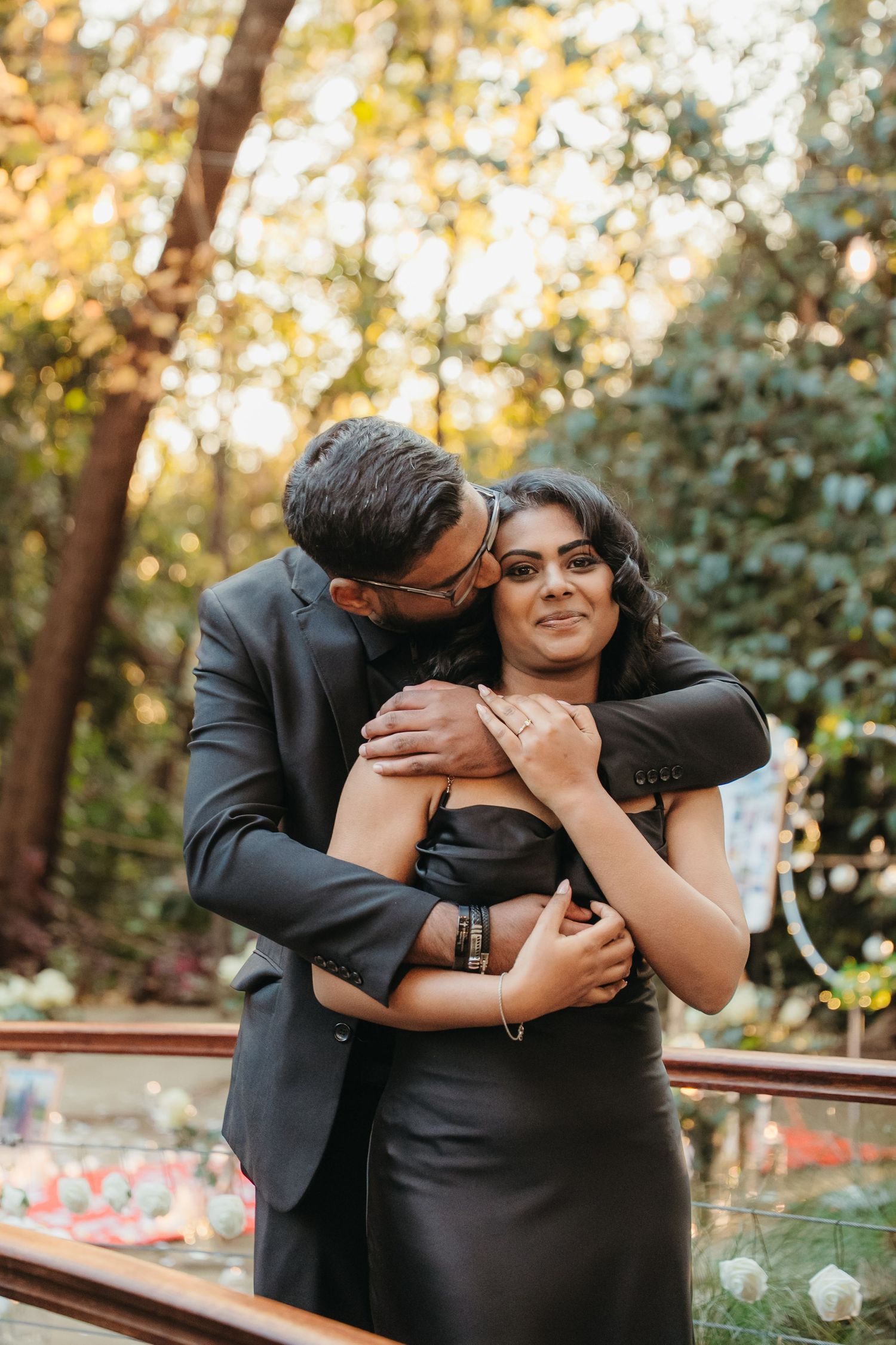 Couple in black formal wear embracing outdoors, man kissing woman's cheek near garden railing.