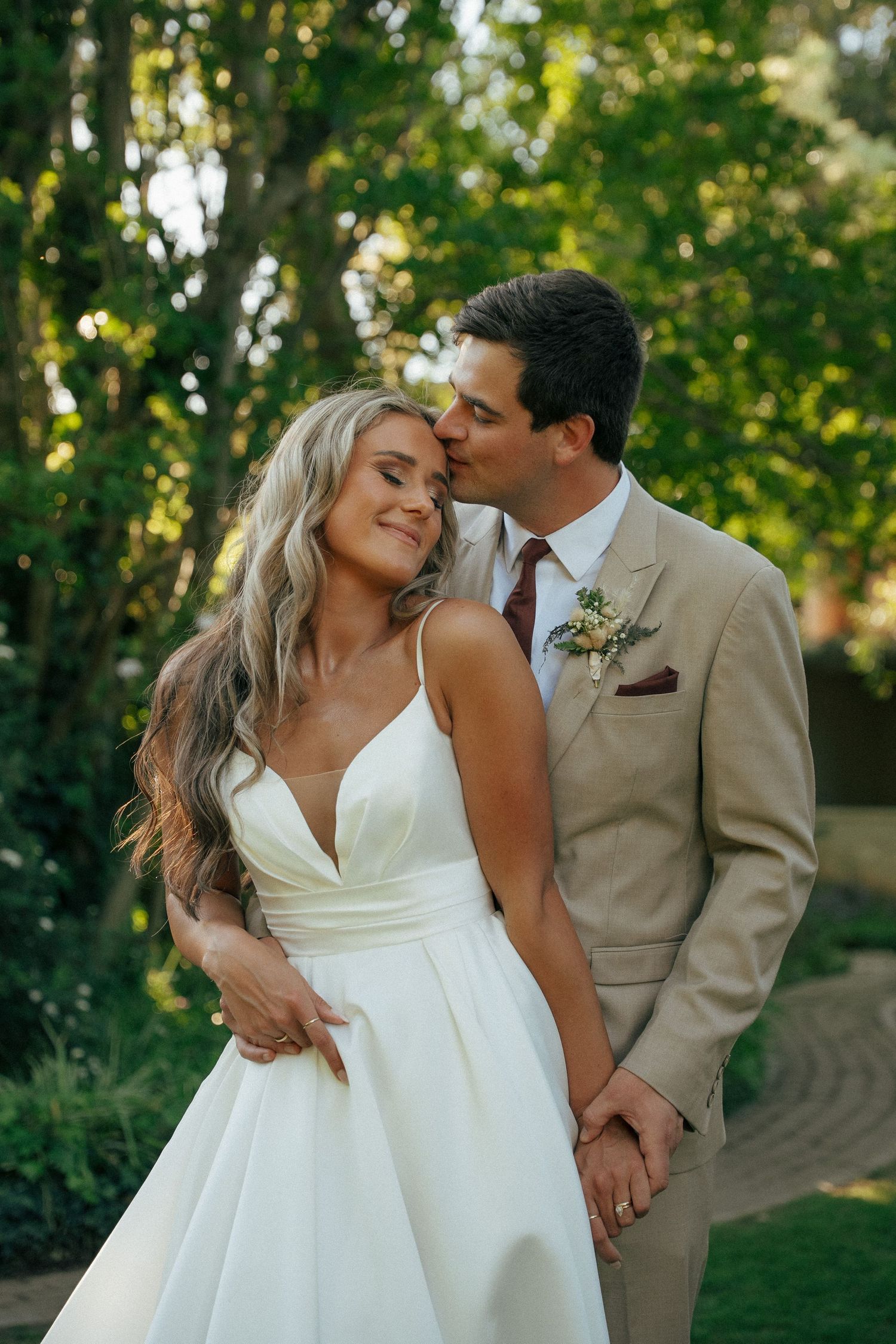 Groom in a tan suit kisses his smiling bride in a white satin wedding dress as they embrace in a sunlit, leafy garden.