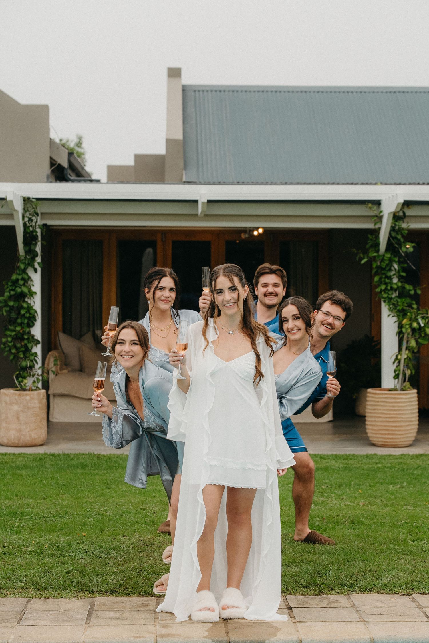 Bride in white dress toasts champagne with bridesmaids and friends outside a modern home in Plettenberg Bay.
