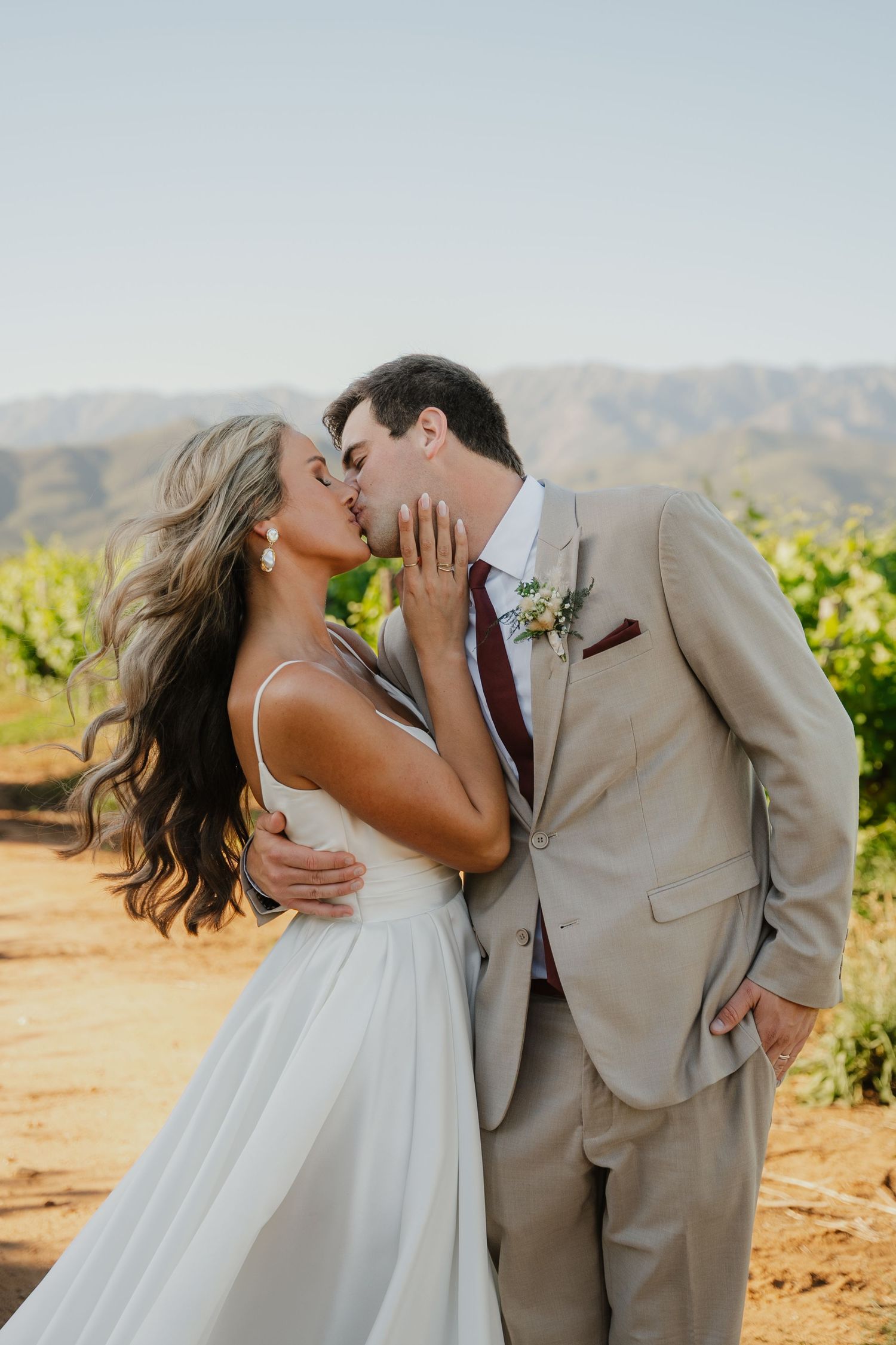 A bride and groom share a romantic kiss at a vineyard wedding, with mountains and lush greenery in the background.