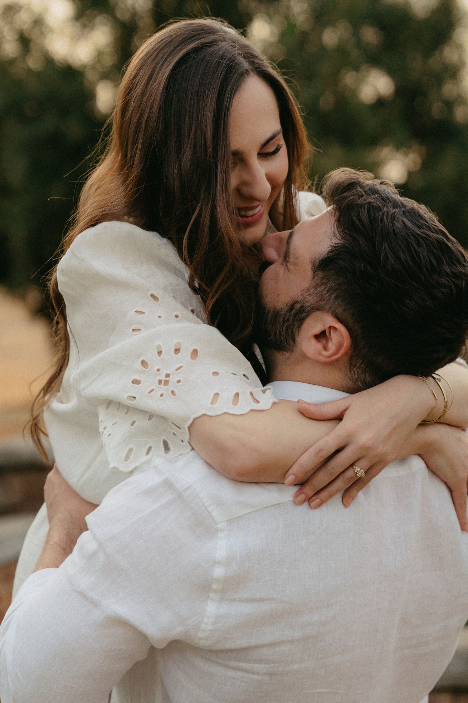 Woman in white lace top laughing while being lifted by man during outdoor engagement photo session.