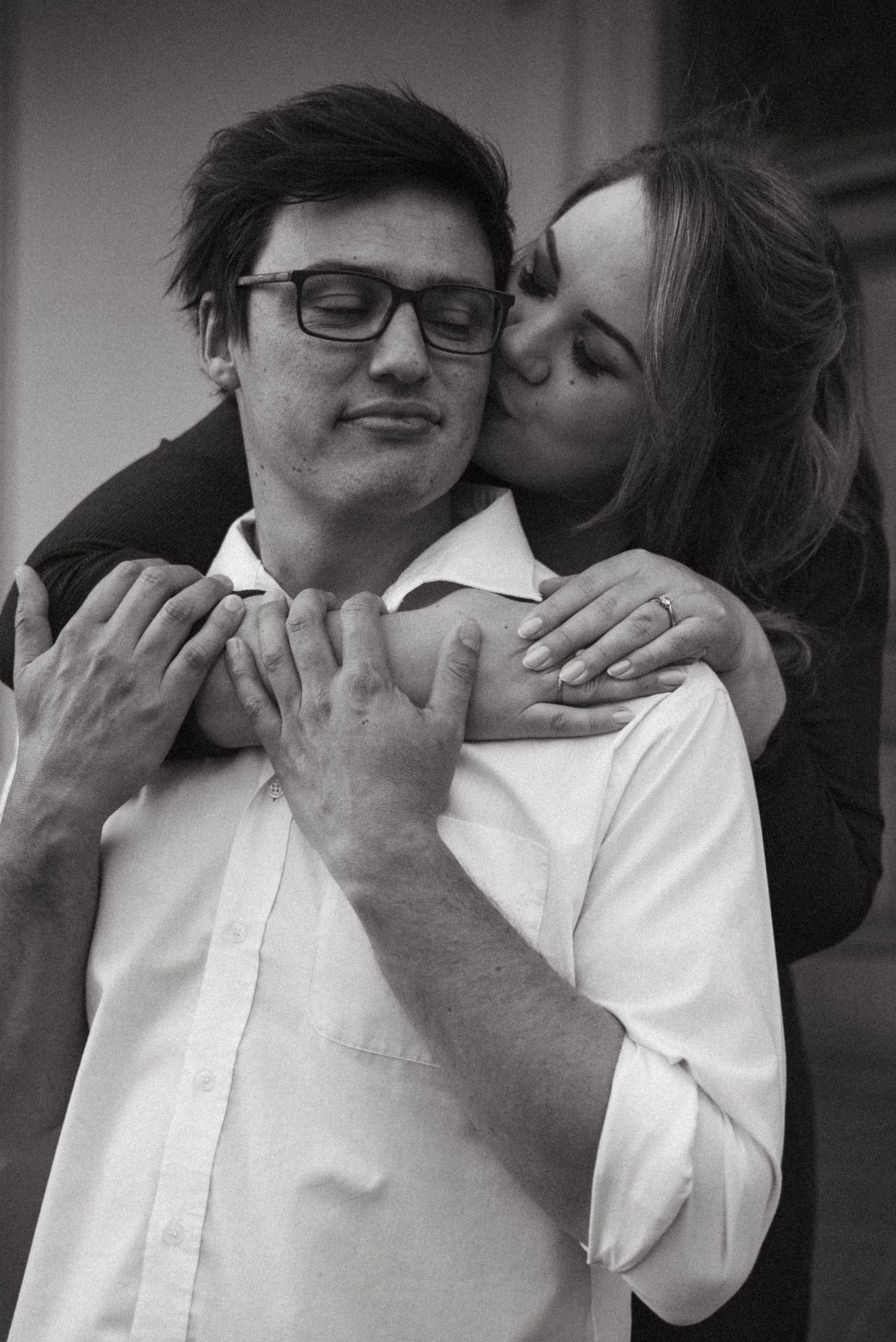 Black and white photo of a woman kissing a man on the cheek while hugging him from behind in Pretoria