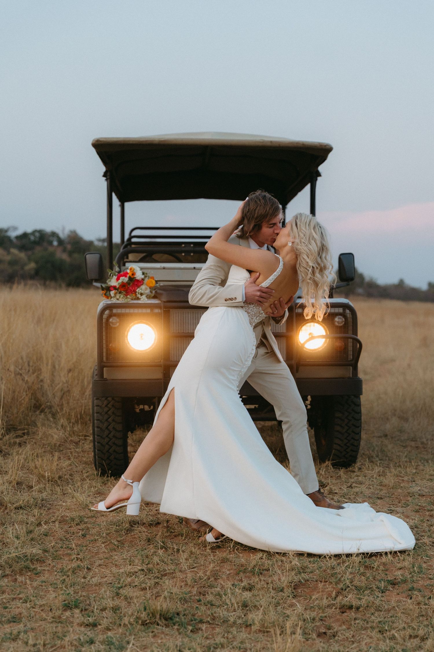 Bride and groom kiss passionately in front of a vintage Land Rover at dusk in Limpopo