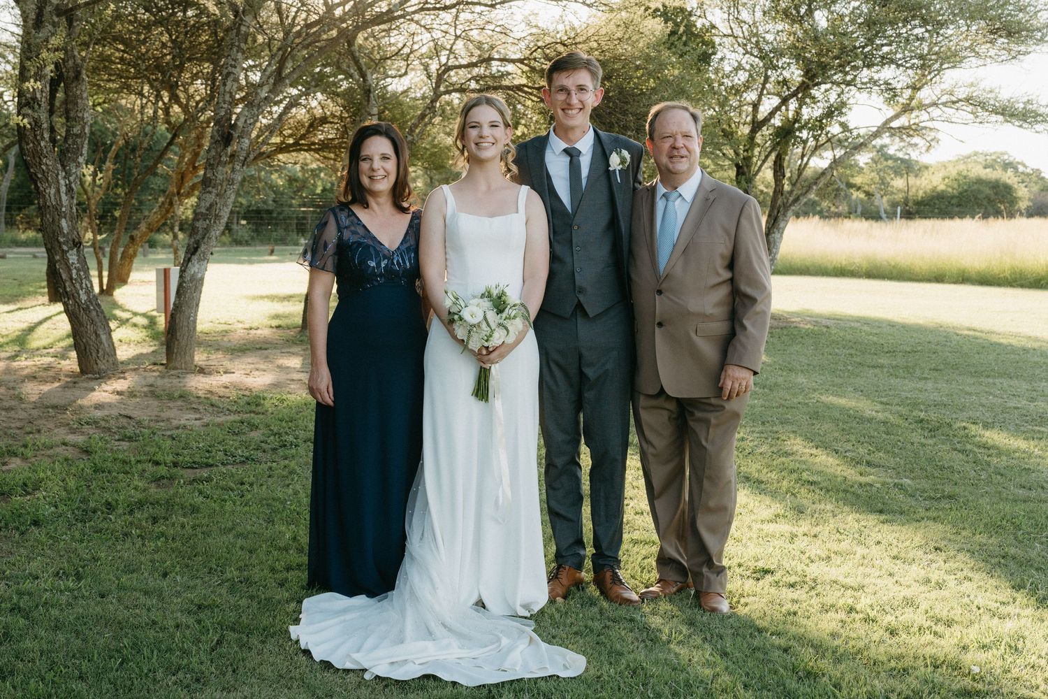 Family formal portrait with wedding party on a sunny lawn with trees in background.