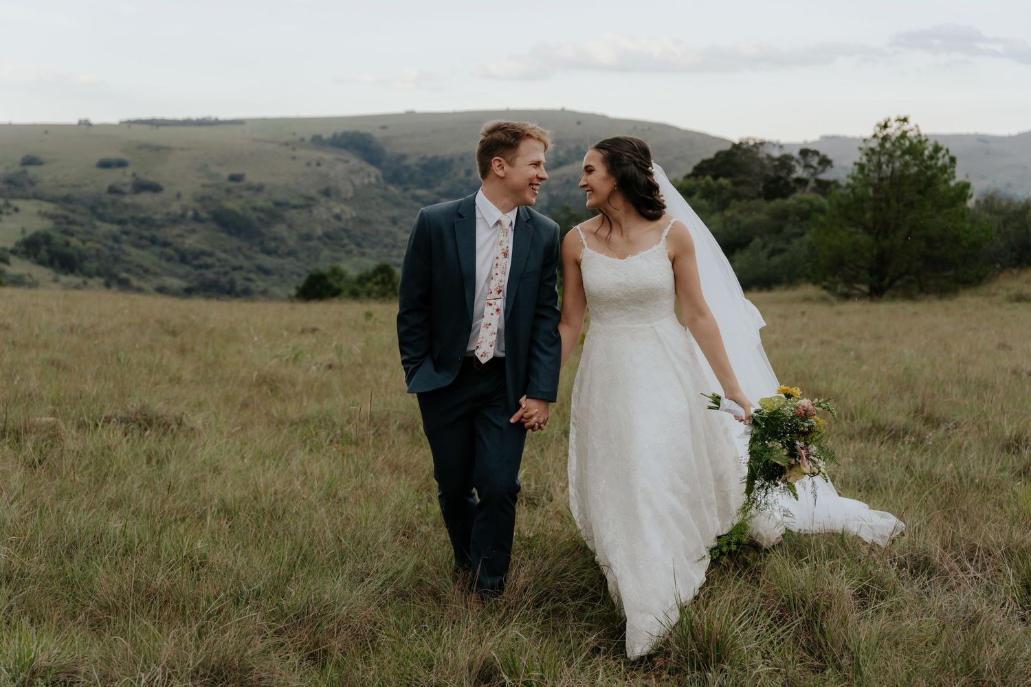 A couple in wedding attire walks together through a scenic hillside meadow with mountains in the background.