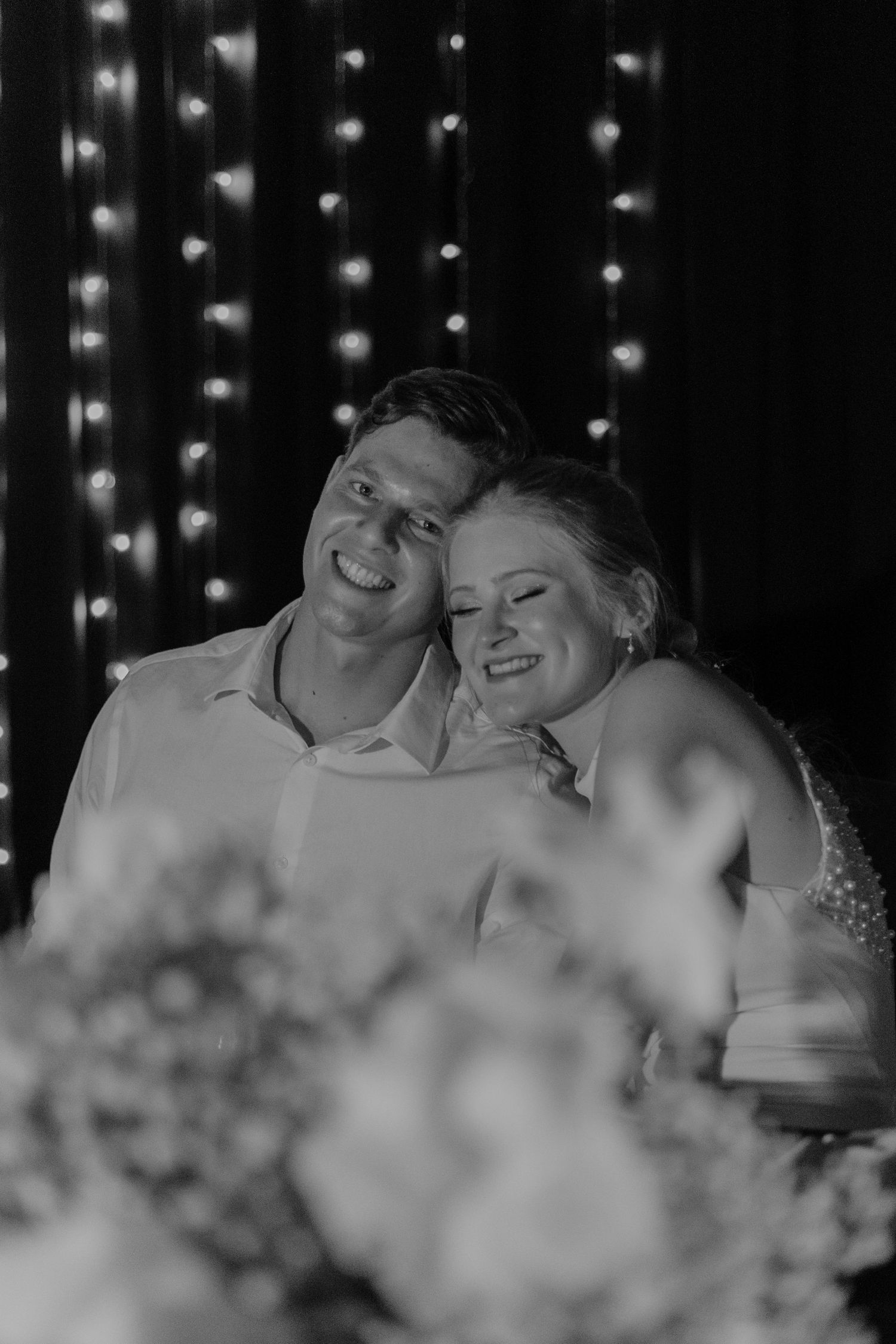Black and white romantic moment captured with twinkling lights in background.