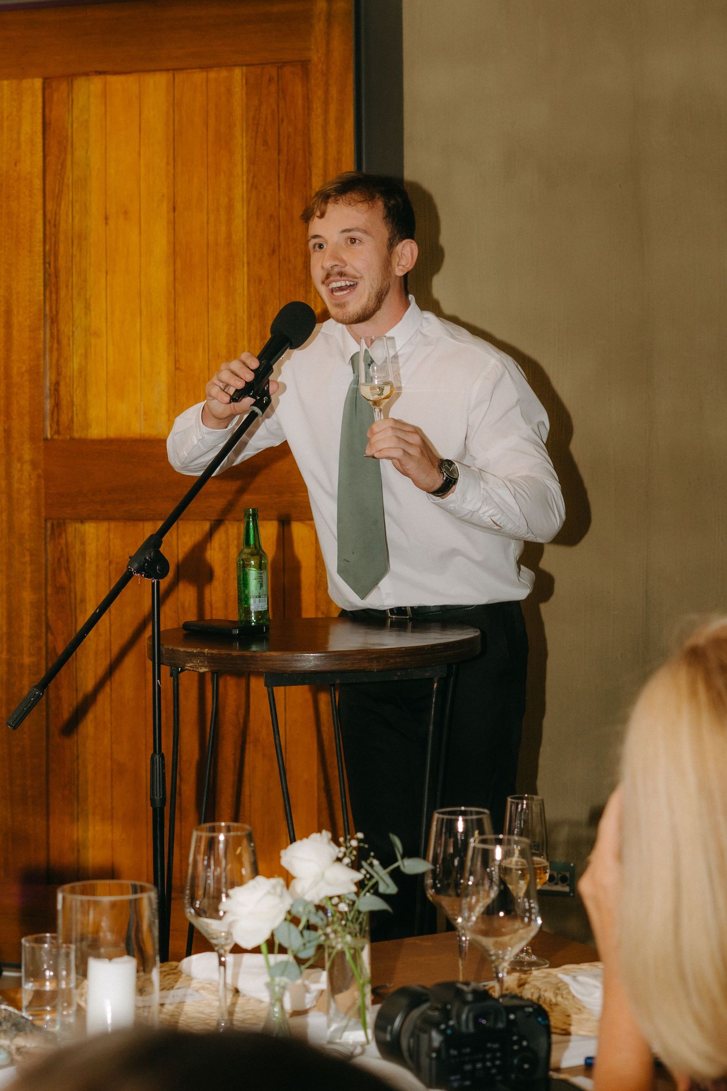 A guest gives an animated wedding toast while holding a microphone and champagne glass.