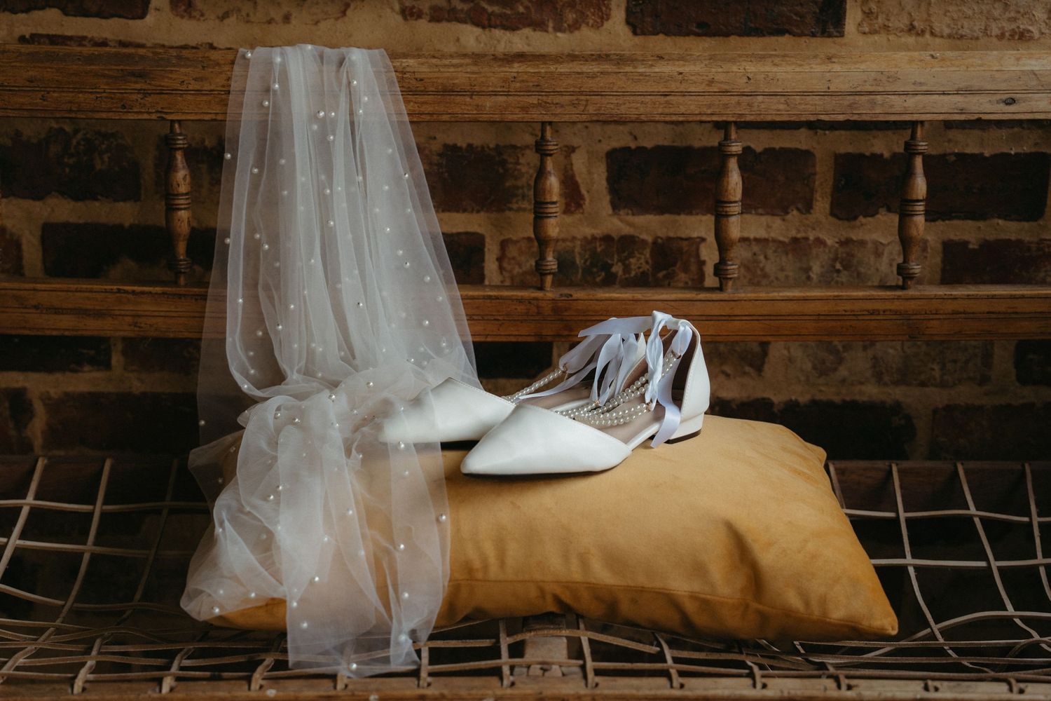 Wedding accessories including a delicate white veil and silver shoes arranged on a golden pillow against brick backdrop.