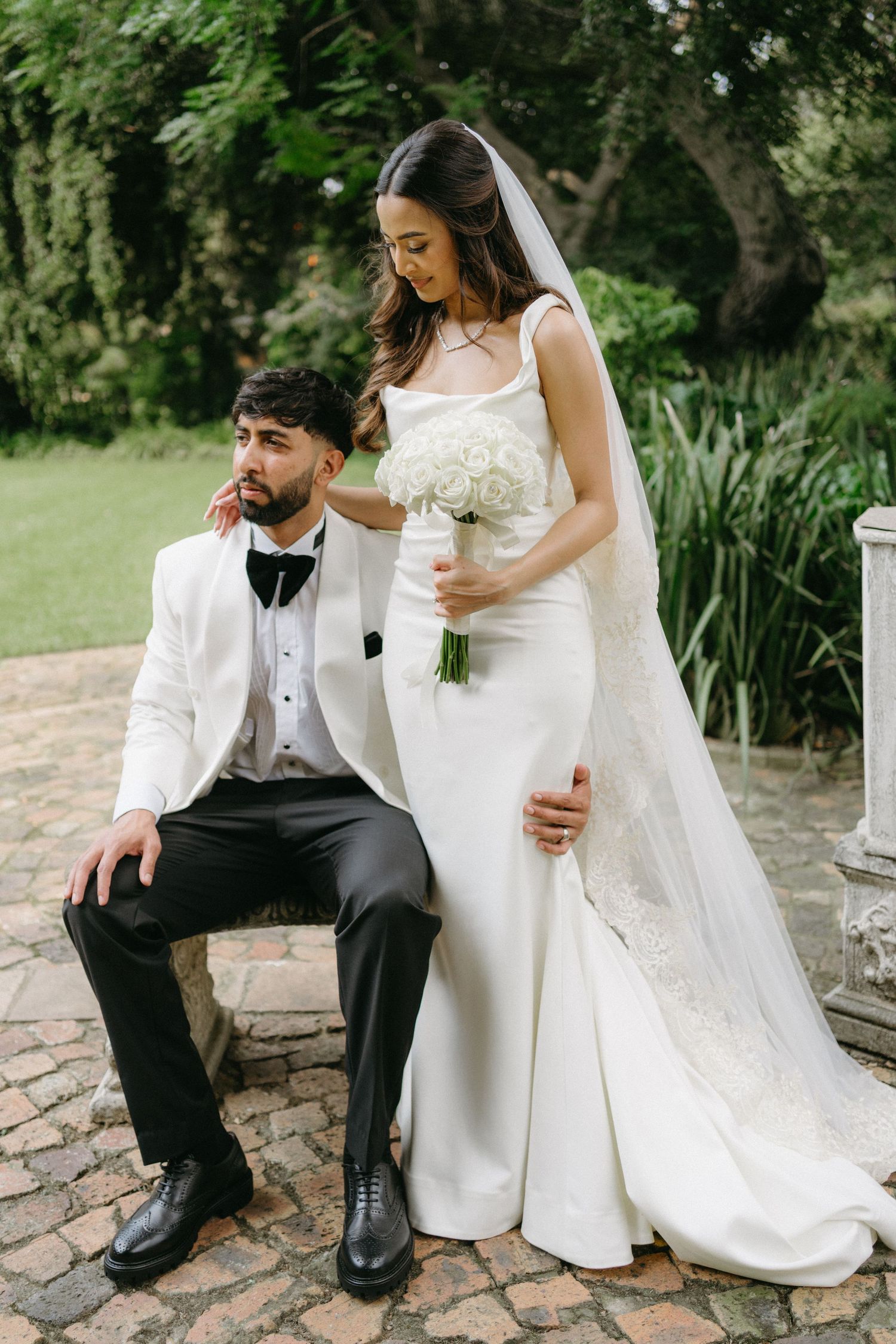 Elegant wedding portrait featuring bride in sleek white gown holding bouquet while groom sits.