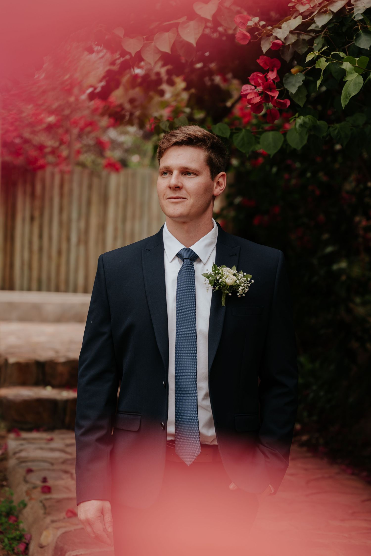 Groom in classic black suit with blue tie stands in front of vibrant pink bougainvillea flowers.