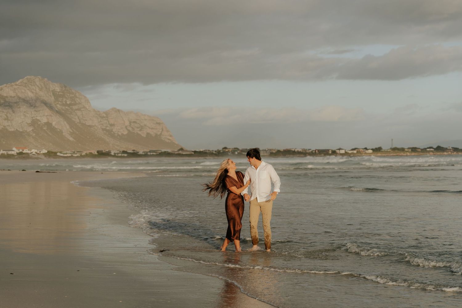 Couple walking hand in hand along a sandy beach at golden hour with mountains and cloudy sky in background.