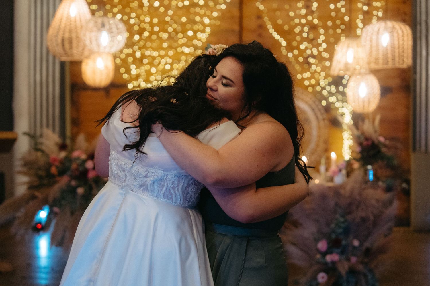 Emotional embrace between bride and bridesmaid at wedding reception with sparkly backdrop.