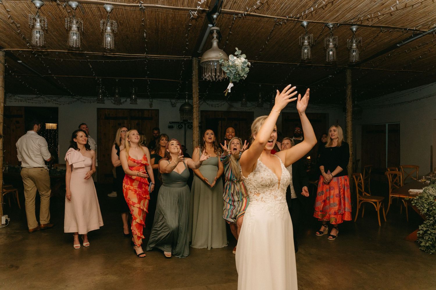 Wedding guests reach for a bouquet being tossed in a rustic indoor venue with string lights.