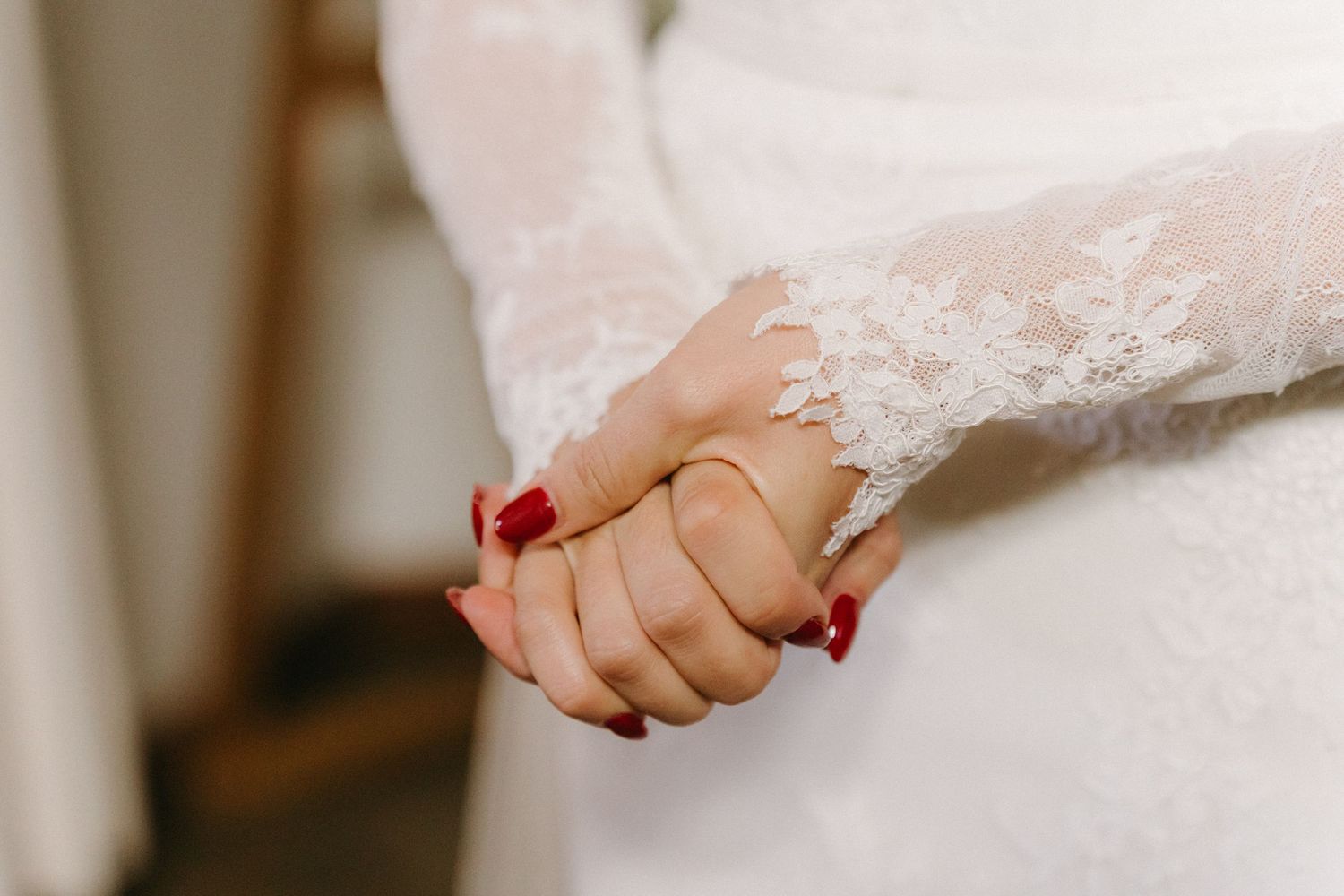 Close-up of clasped hands showing red nails against white lace wedding dress fabric.