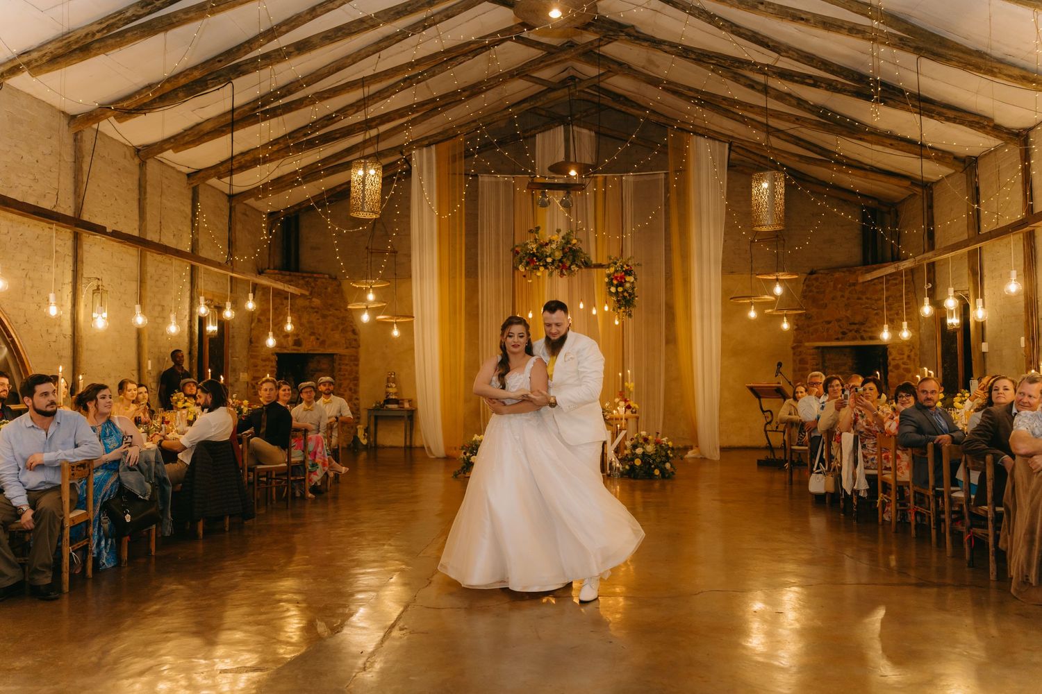 Newlyweds share their first dance in an elegant wood-beamed reception hall with seated guests.